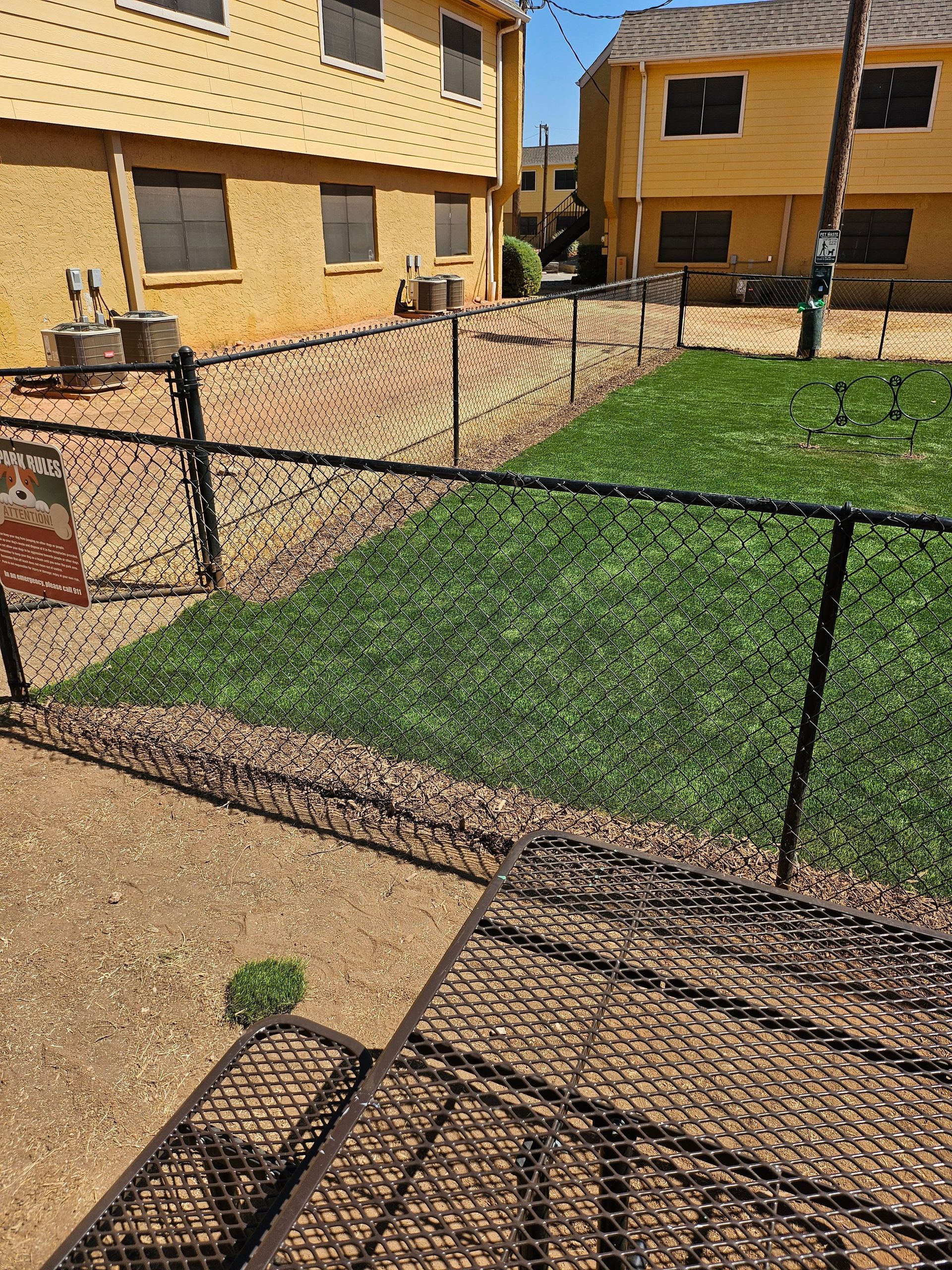 Chain-link fence separates grass from gravel area with buildings in the background.