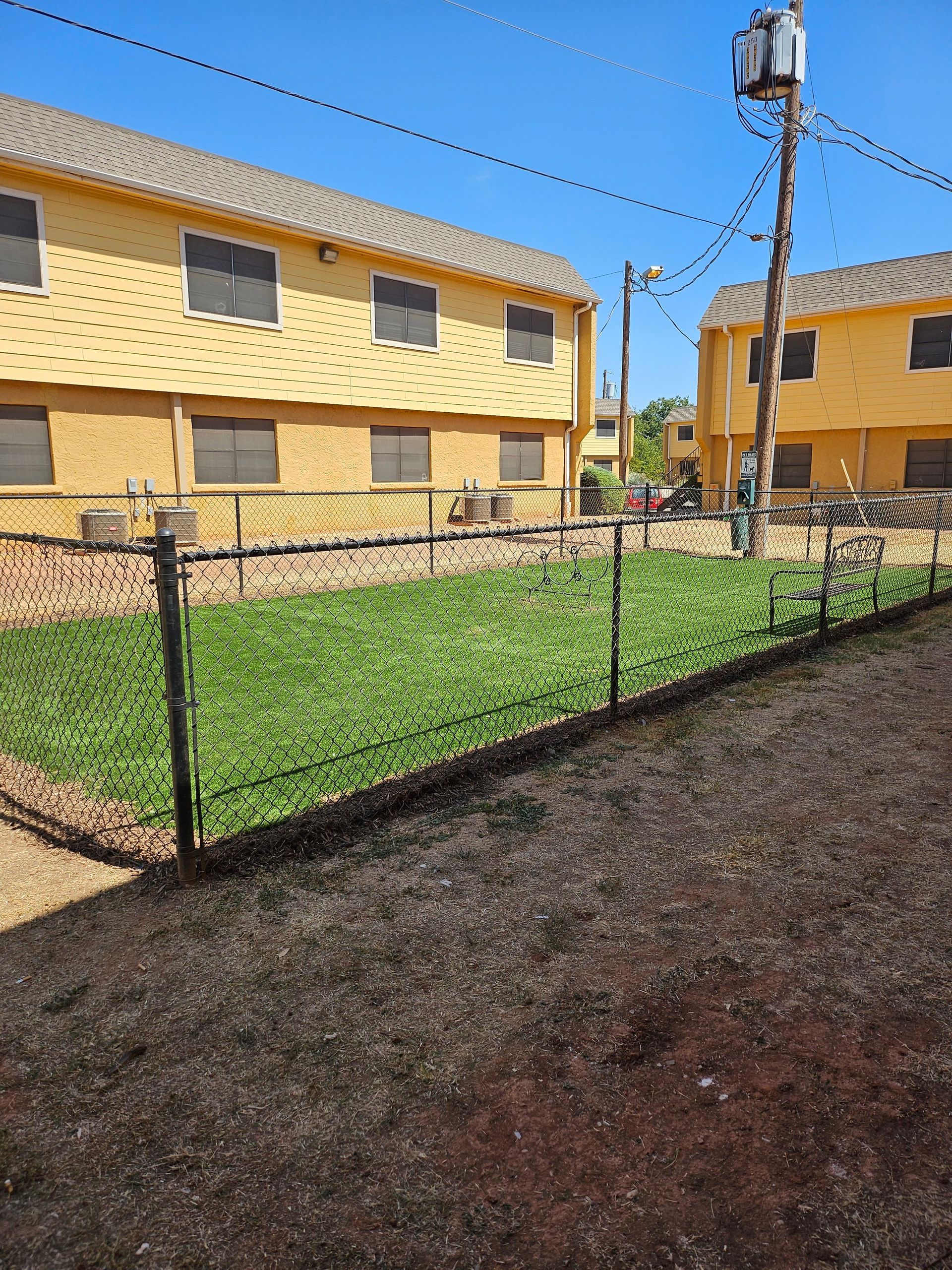 Yellow apartment buildings with a small fenced-in grassy area, power lines, and a utility pole under a blue sky.