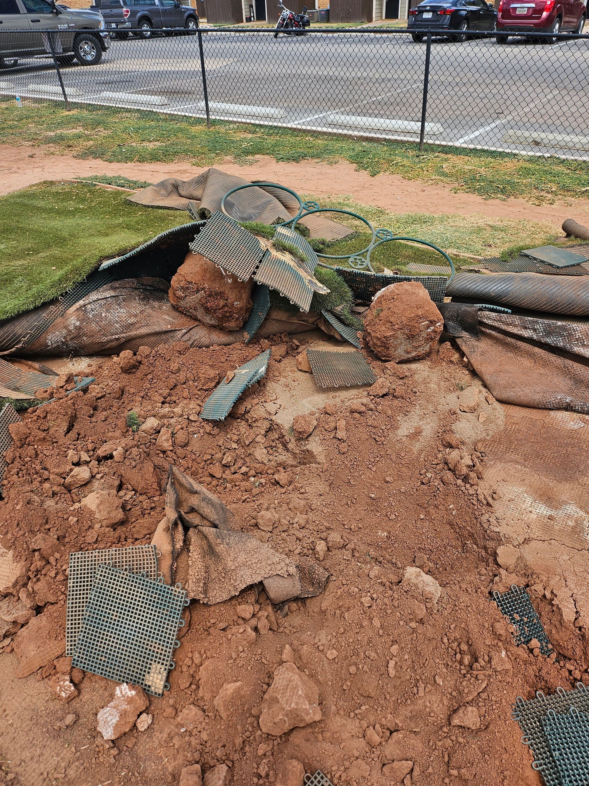 Pile of red dirt with torn artificial turf and netting near a fence and parked cars.