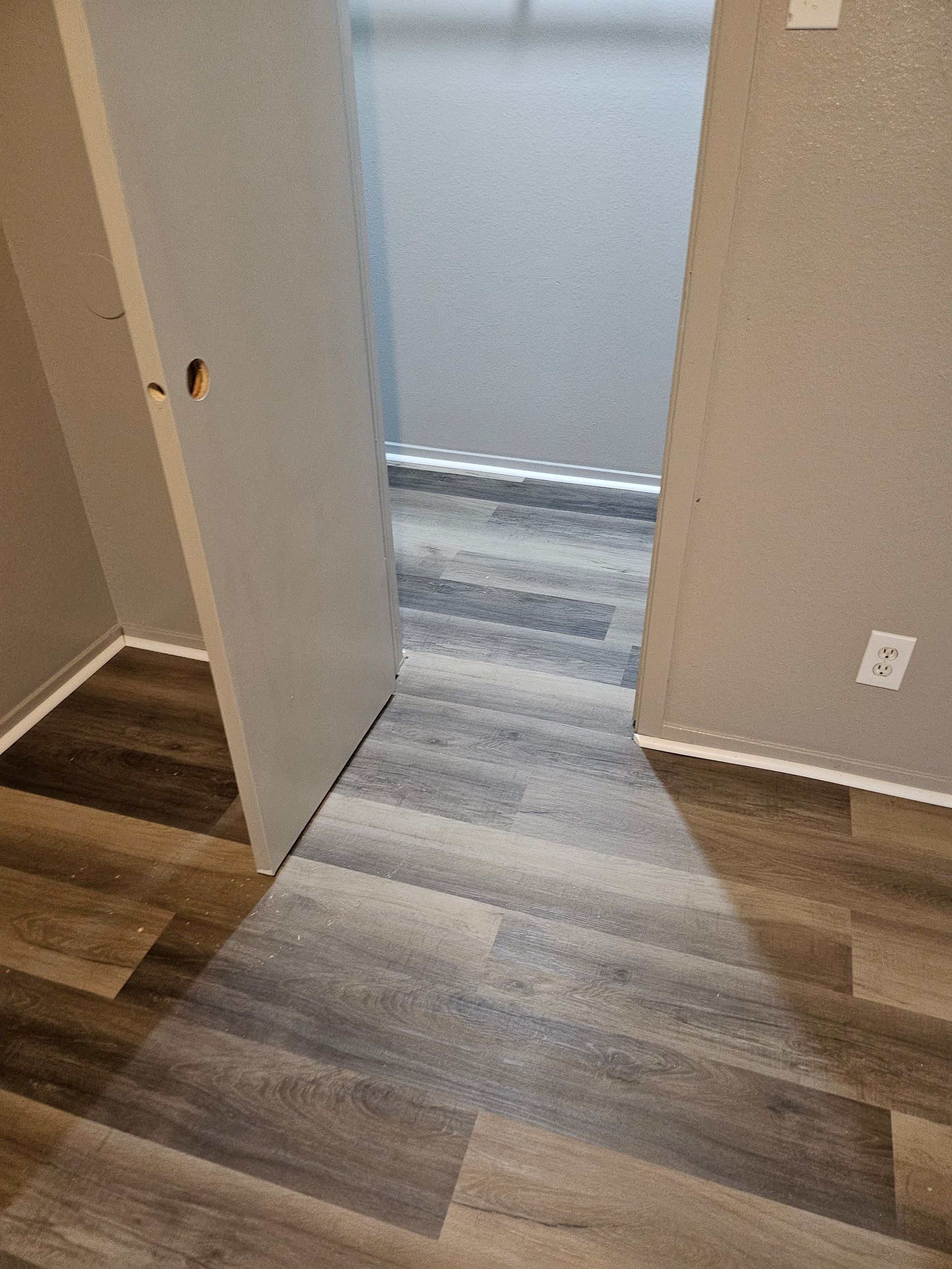 Doorway to a room with grey walls and blue-grey flooring, adjacent to a room with wood-look flooring.