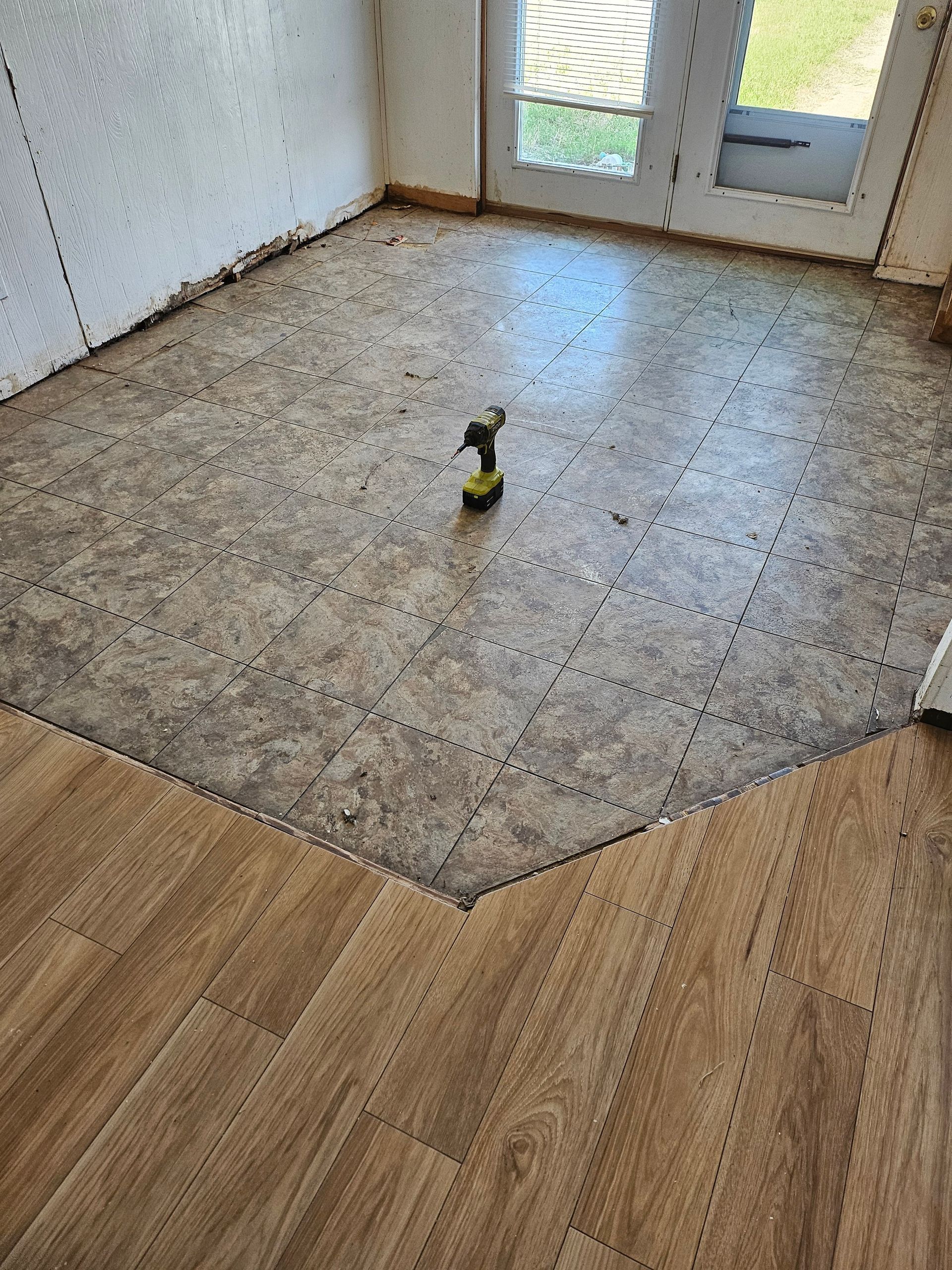 Flooring installation in progress: brown planks partially installed, beige tile section. Drill on floor near door.
