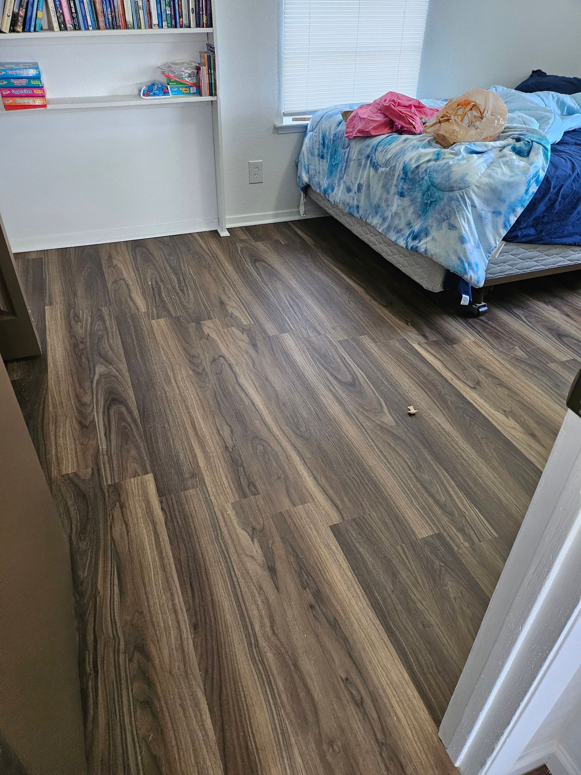 Room with wood-look flooring, bed with blue bedding, and a bookshelf with items on the wall.