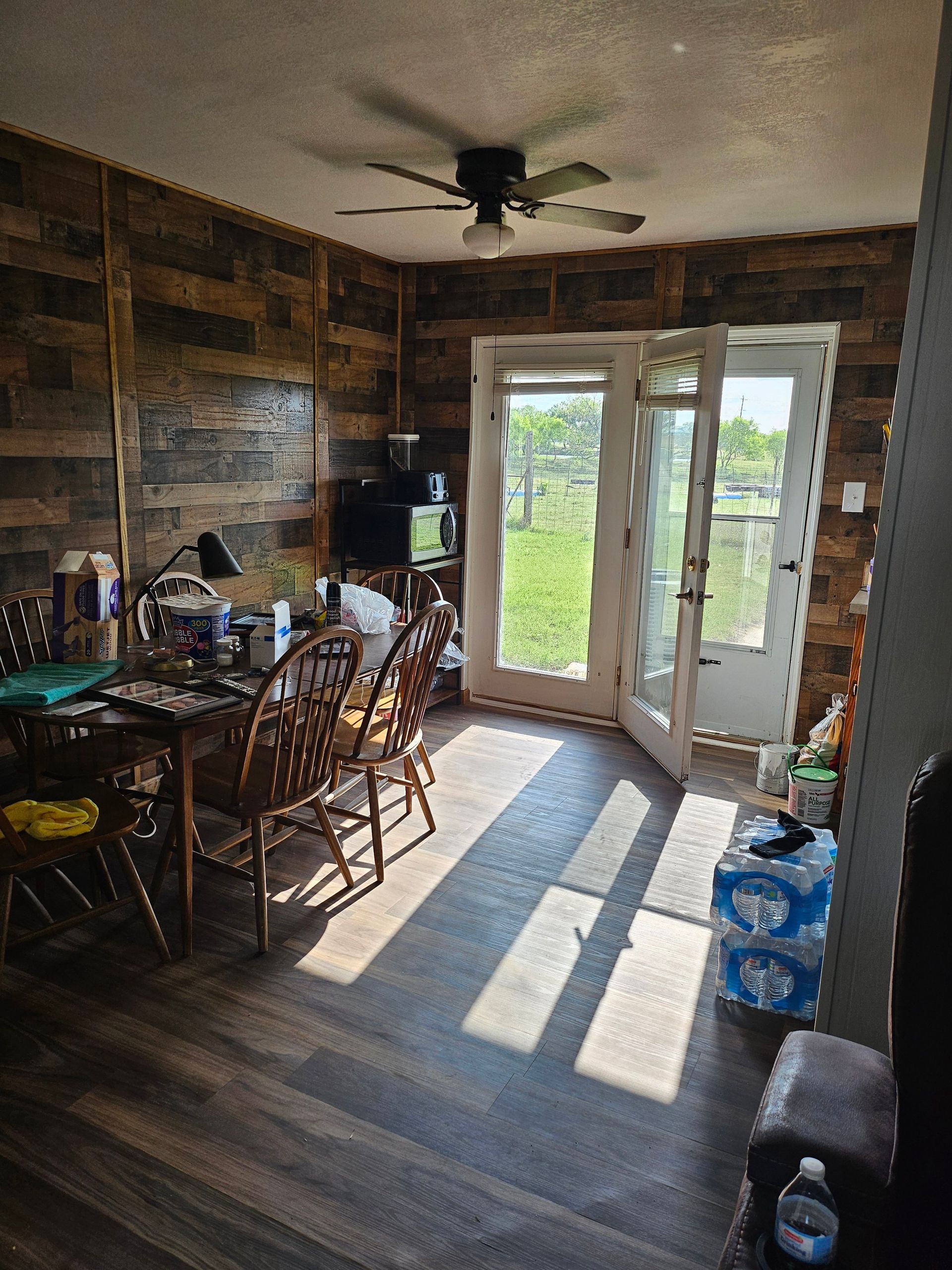 Dining area with wood-paneled walls, table, chairs, and French doors leading outside; sunlight streams in.