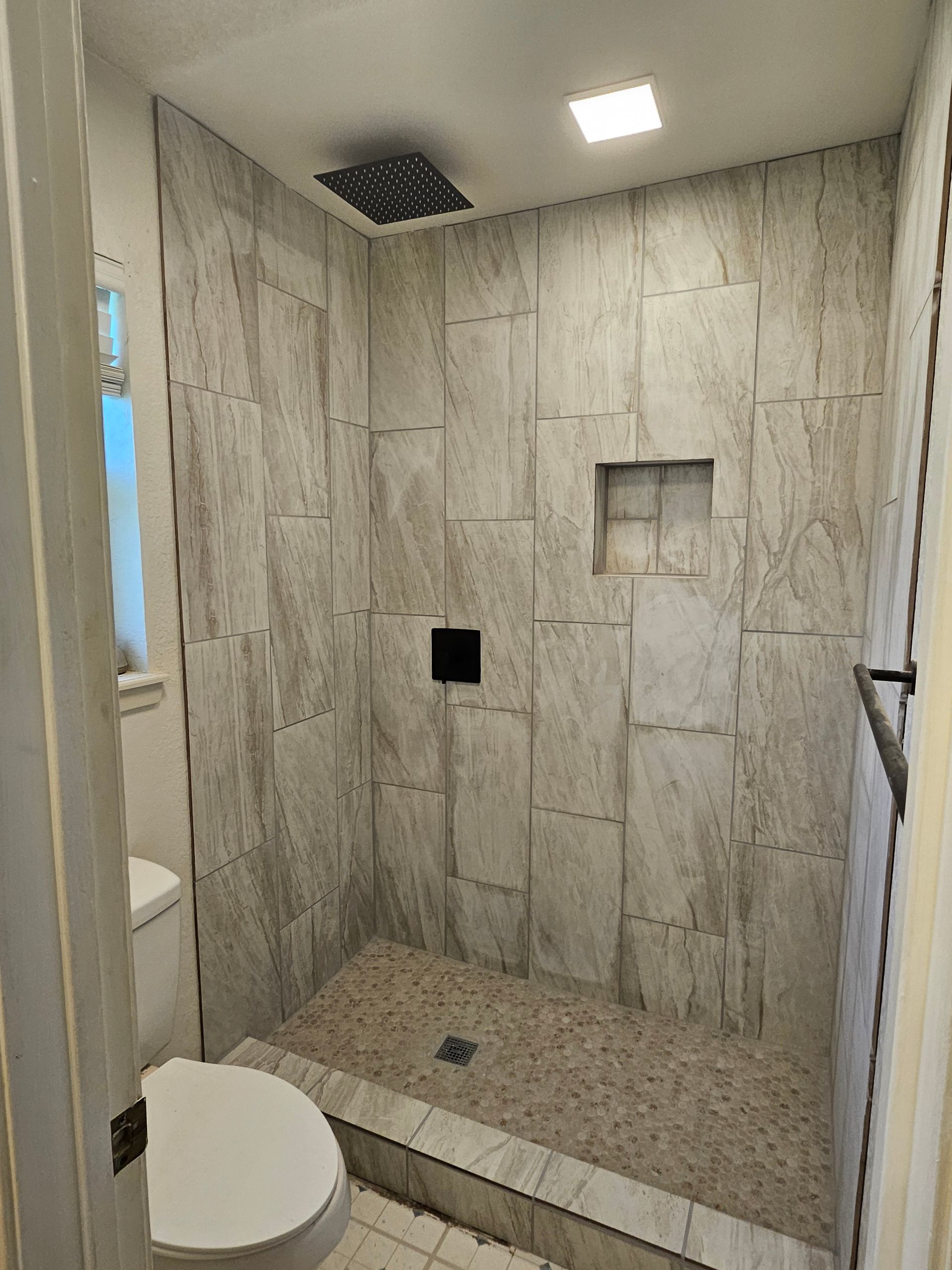 Bathroom with tile shower, pebble floor, built-in shelf, and rain showerhead. Toilet in foreground.