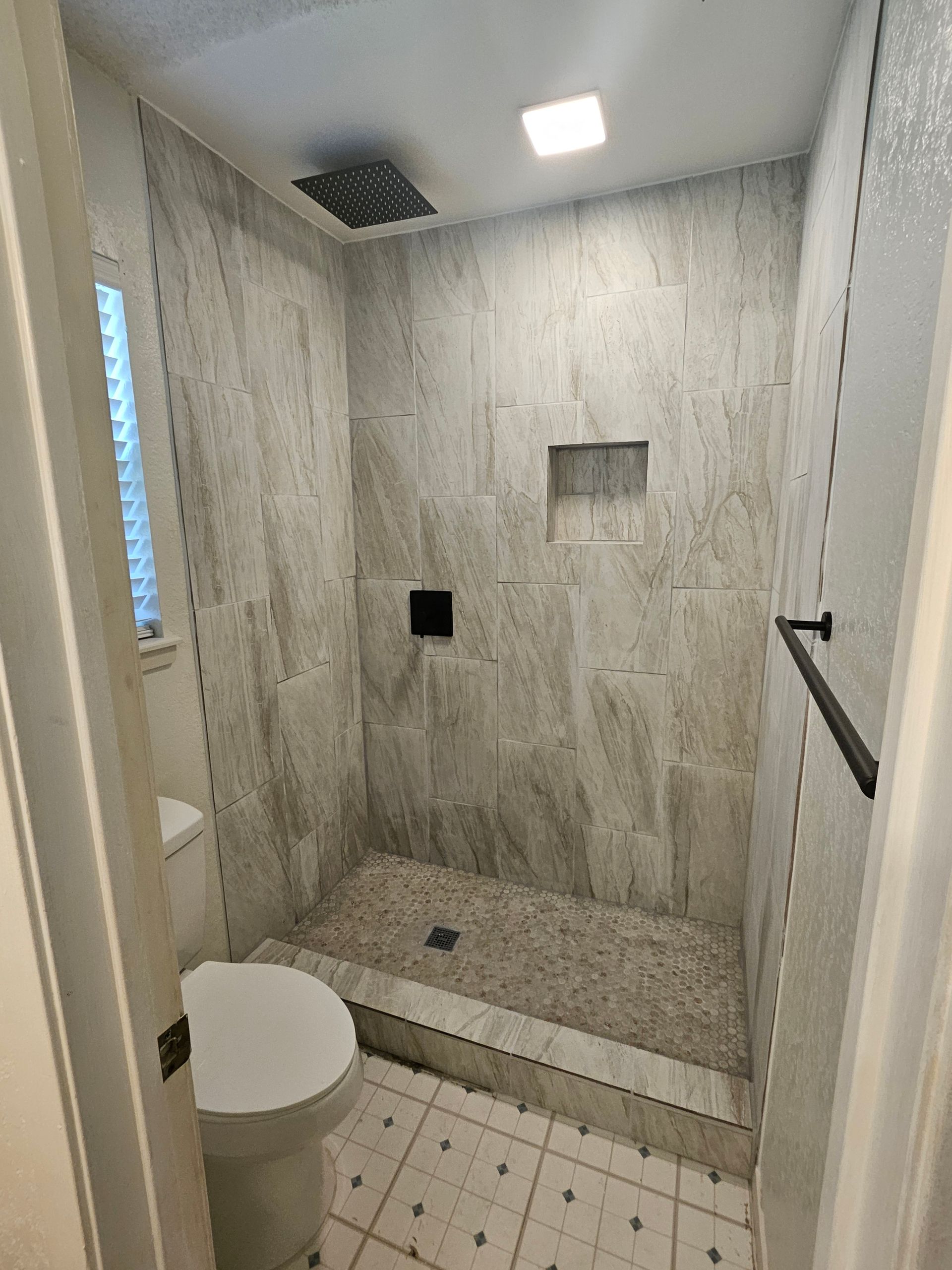 Small bathroom with gray tiled shower and pebble floor, toilet, and black towel bar.