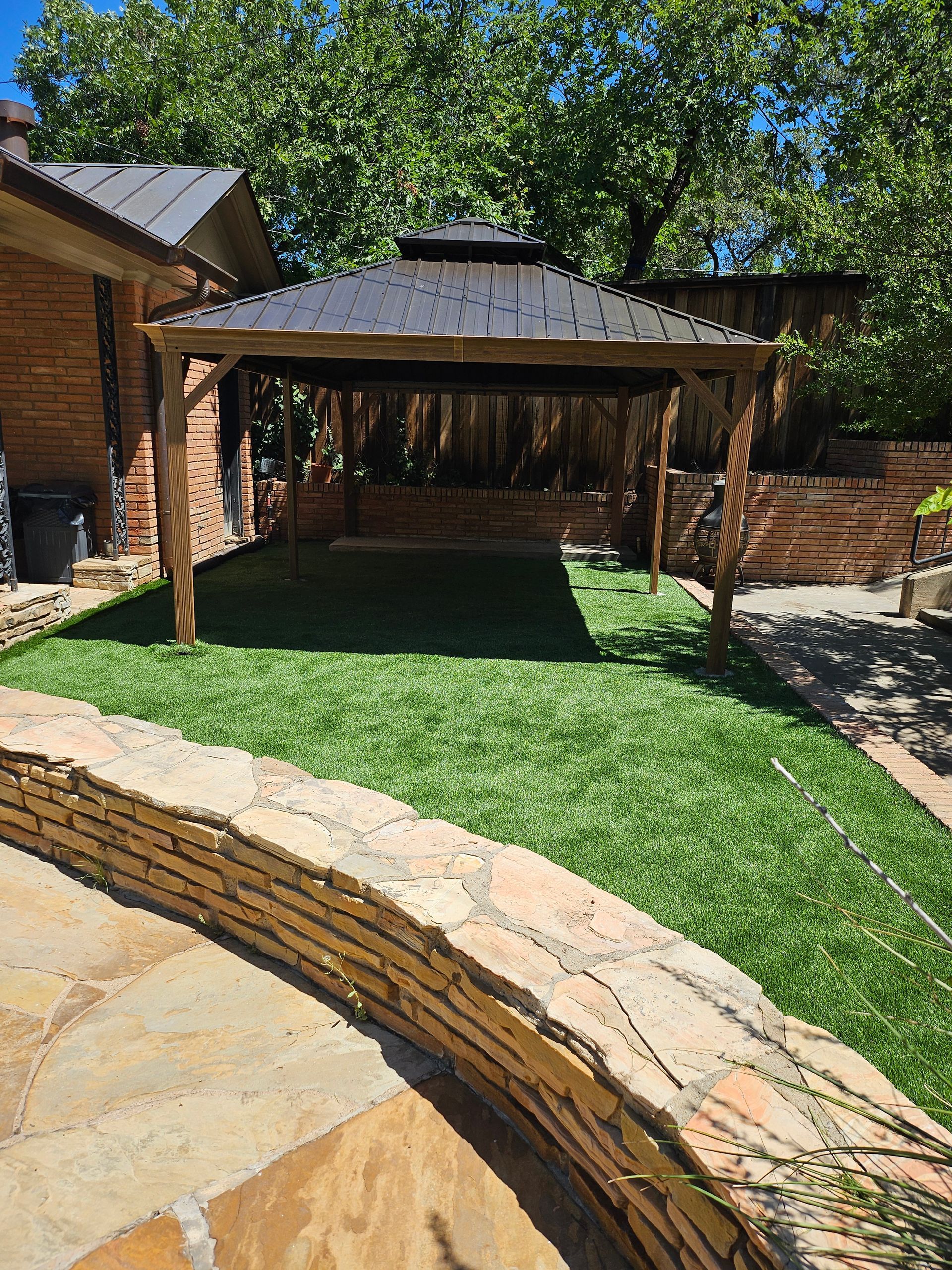 Gazebo on artificial turf in a backyard, surrounded by a stone wall and brick structure.