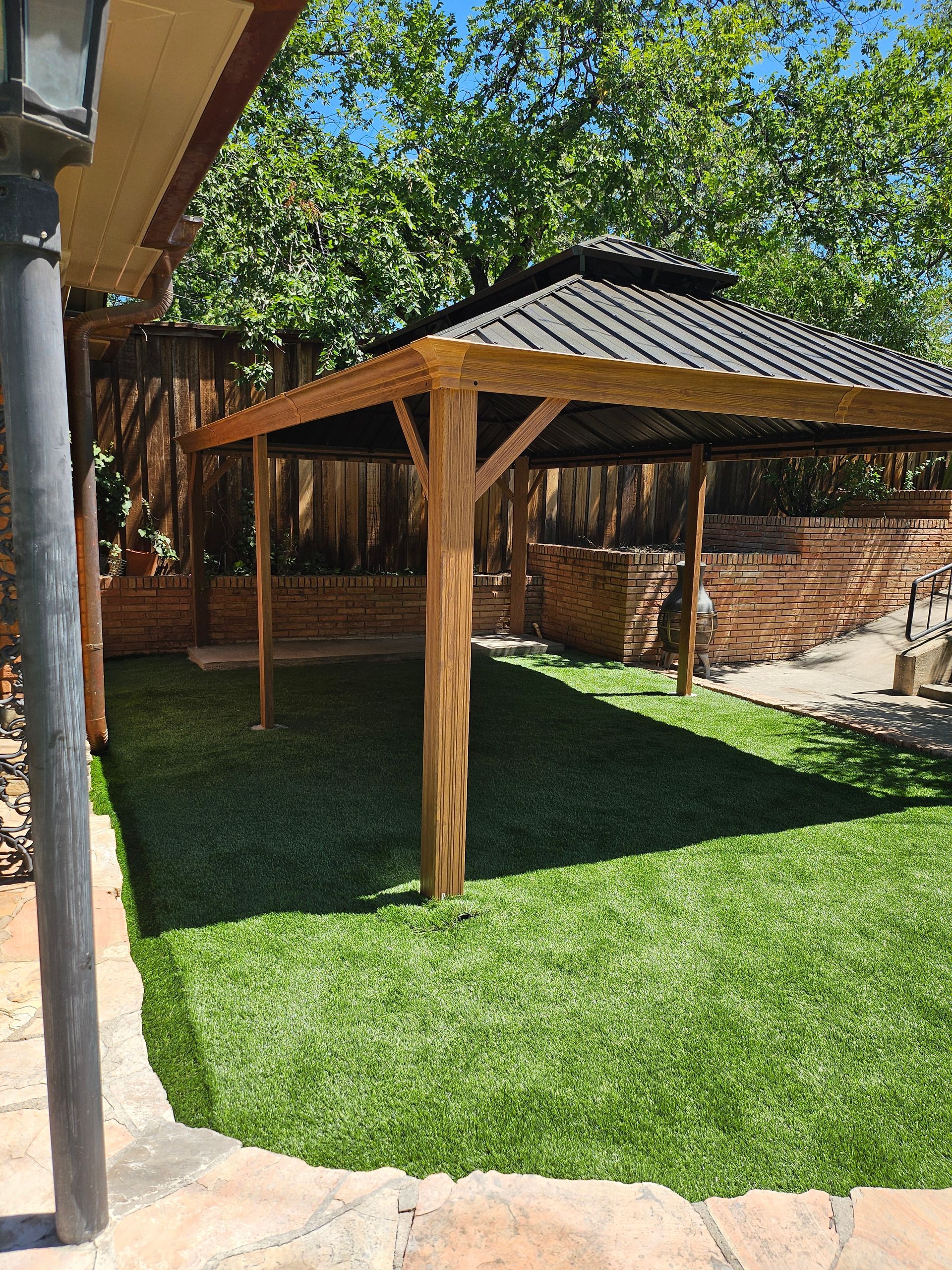 Wooden pergola with dark roof over a patch of artificial green grass, surrounded by brick and stone.
