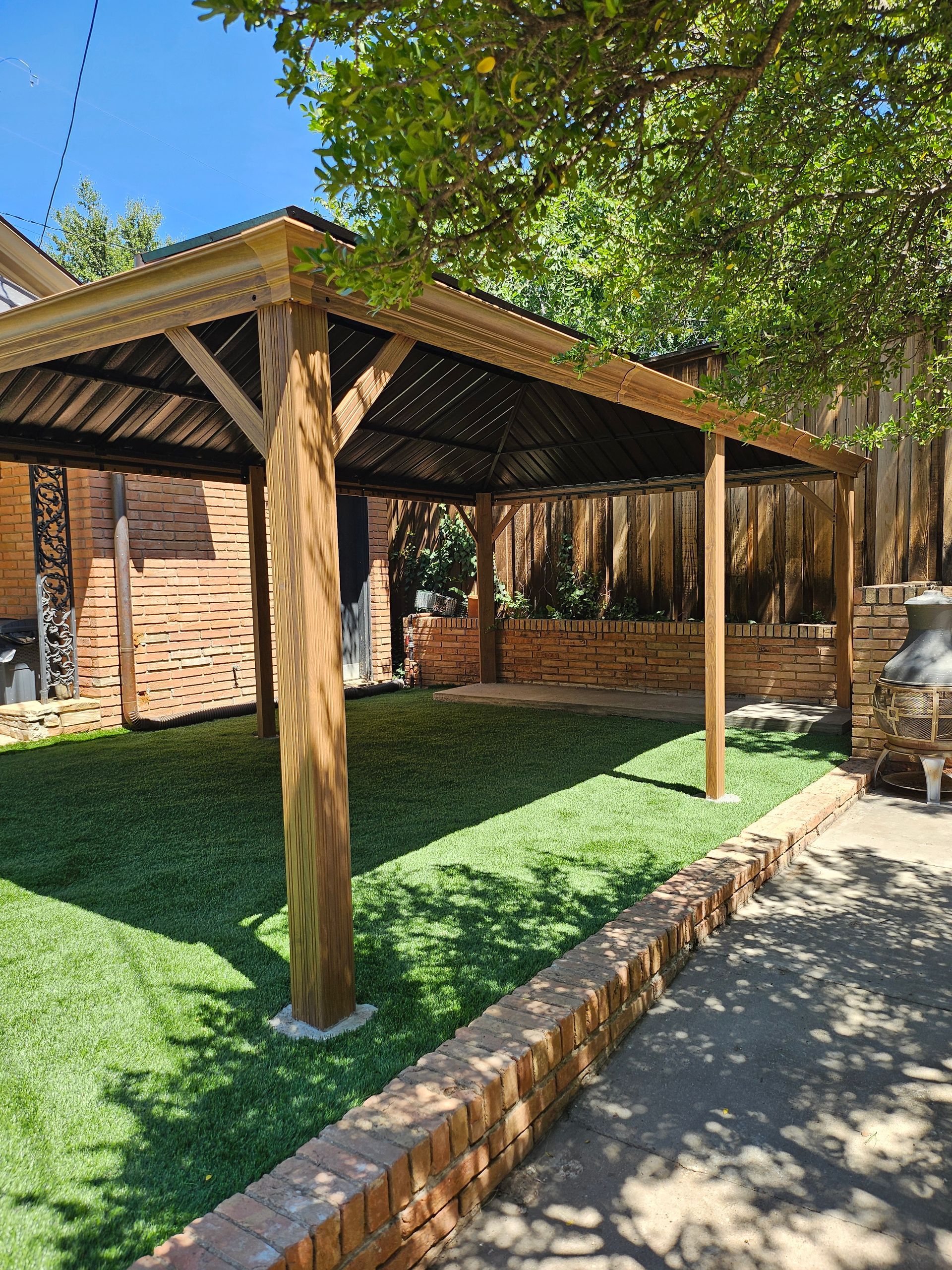 Wooden pergola in a backyard with green turf, brick walls, and a tree on a sunny day.