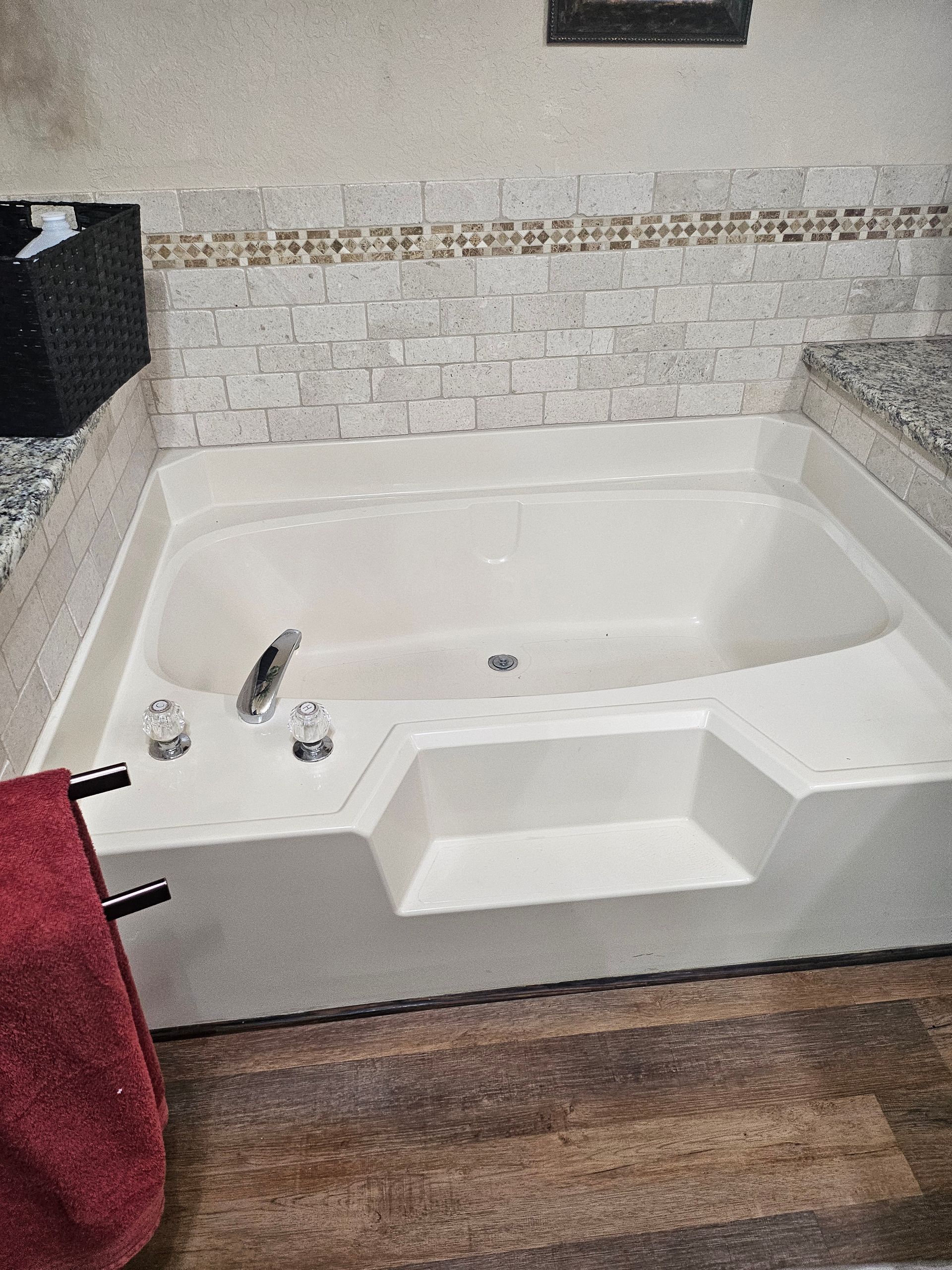 Built-in bathtub with step, set against tile and wood-look flooring. Red towel on a nearby bar.