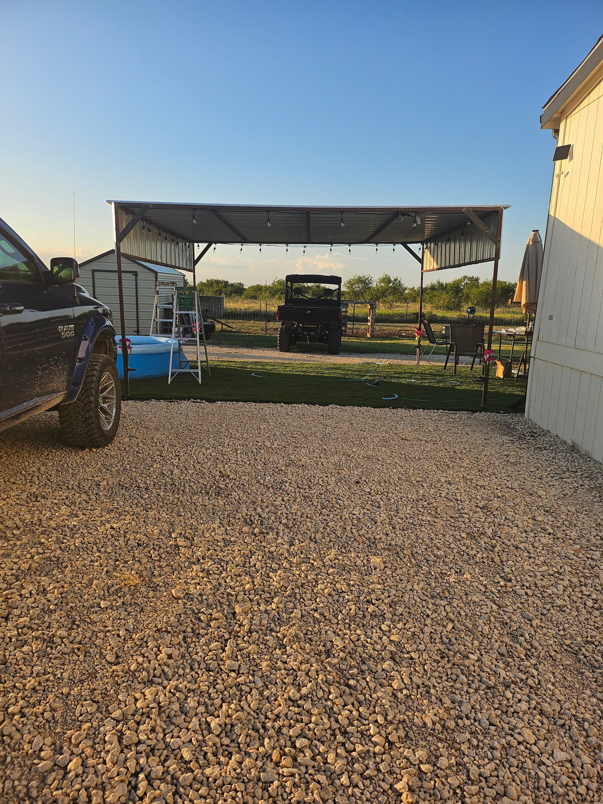 Gravel driveway leads to a shed with a vintage car, garden, and a building on a sunny day.