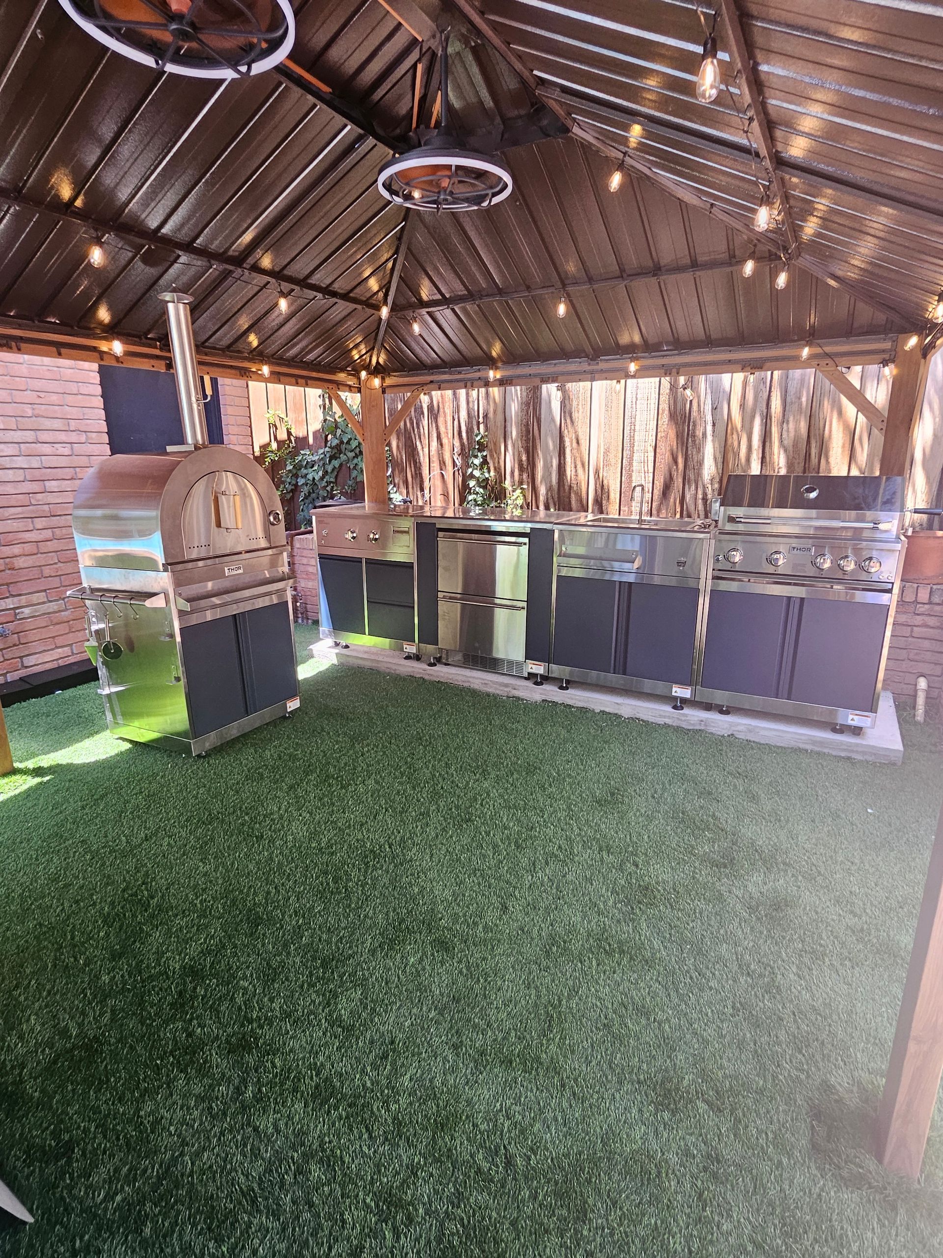 Outdoor kitchen under a wooden pergola with pizza oven, grills, and artificial turf.