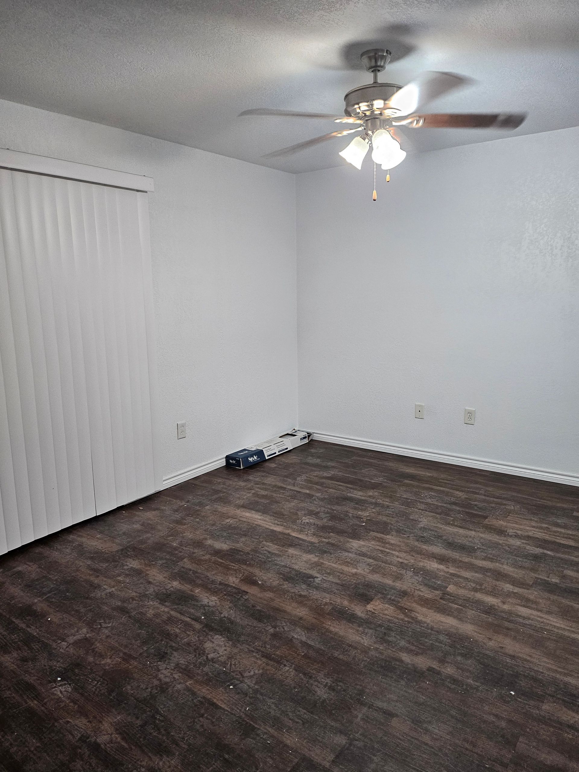 Empty room with dark wood-look flooring, white walls, vertical blinds, and a ceiling fan.