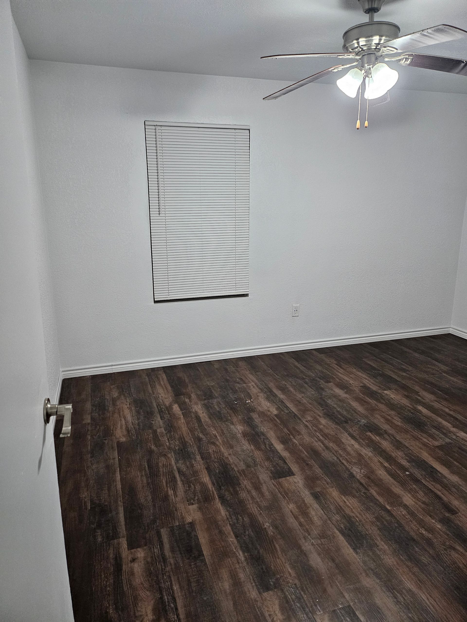 Empty room with dark wood-look flooring, white walls, blinds on a window, and a ceiling fan.