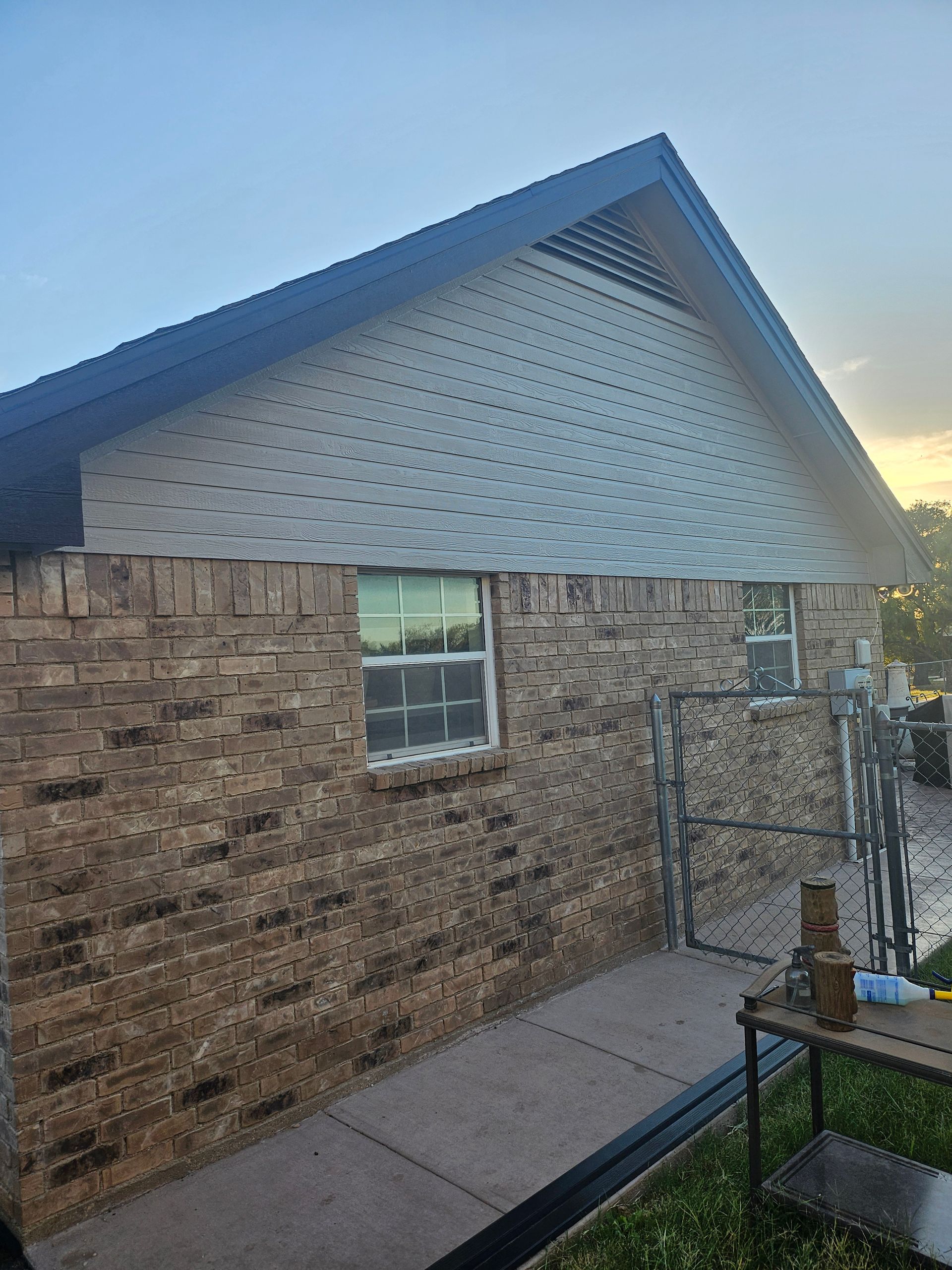 Brick building exterior with a light-colored gable and two windows. A walkway and fence are visible.