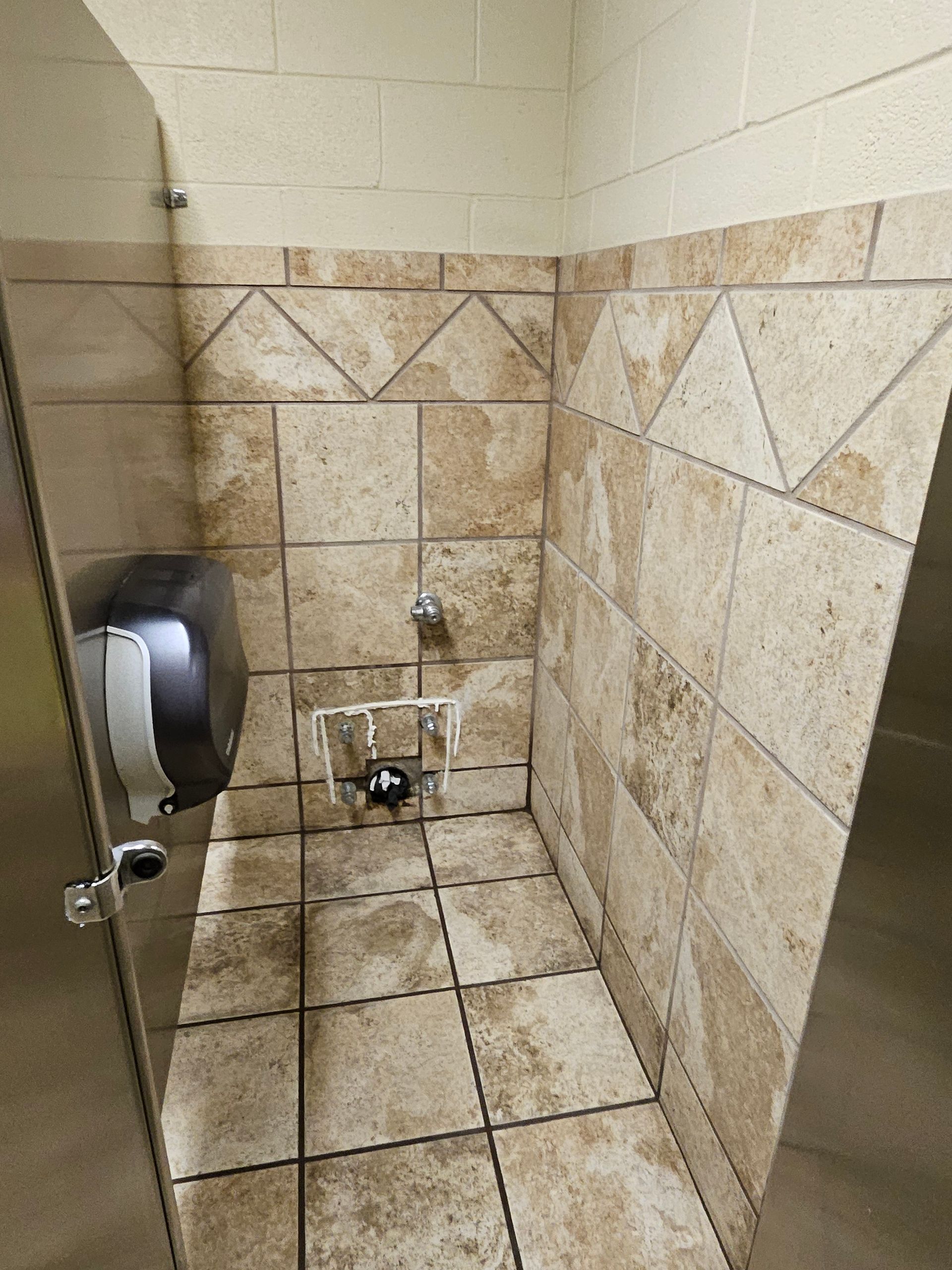 A public restroom stall with light brown tiled walls and floor, a stainless steel door, and a paper towel dispenser.