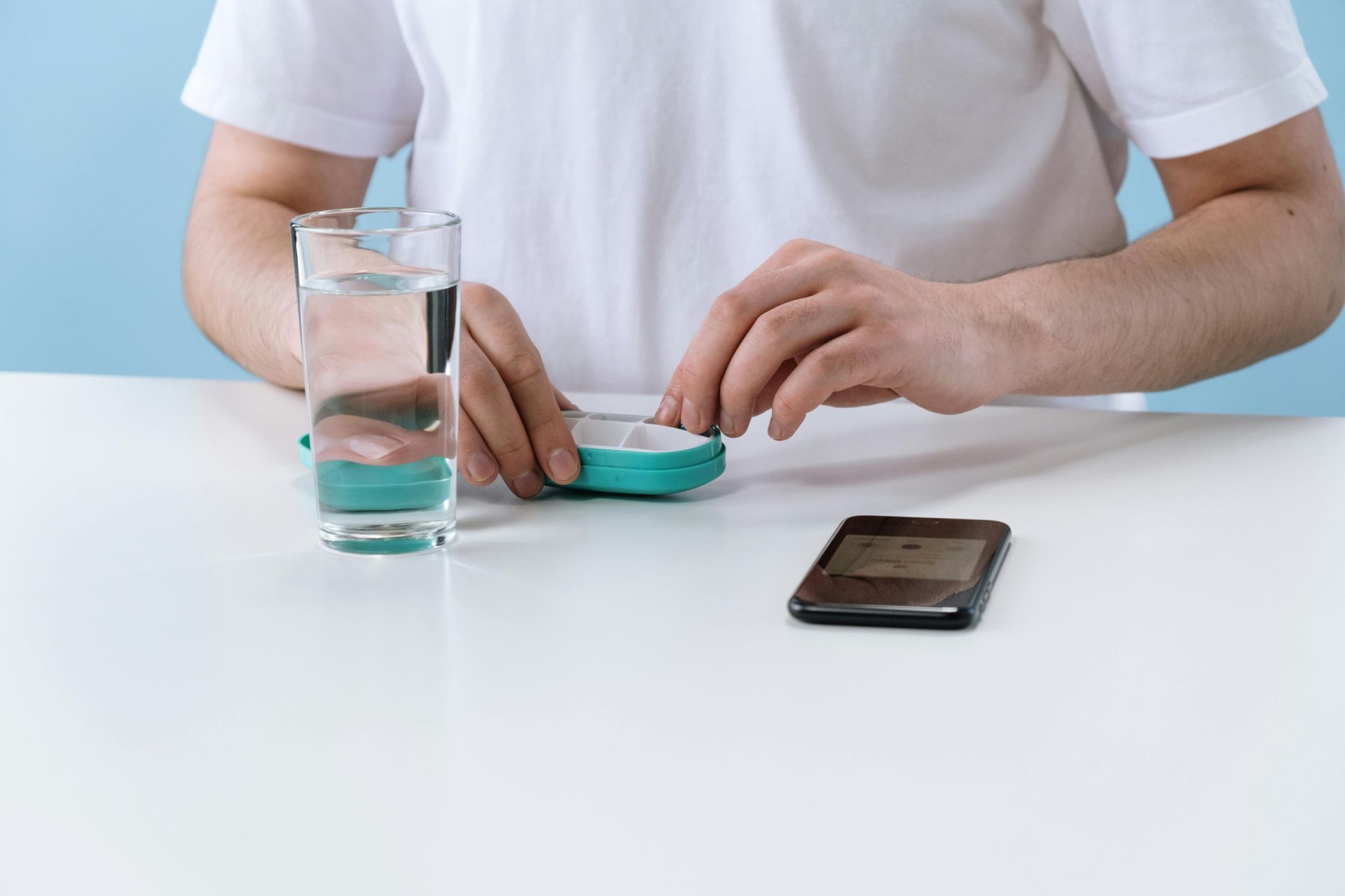 Person preparing medication from a pill organizer, next to a glass of water and phone on a white table.