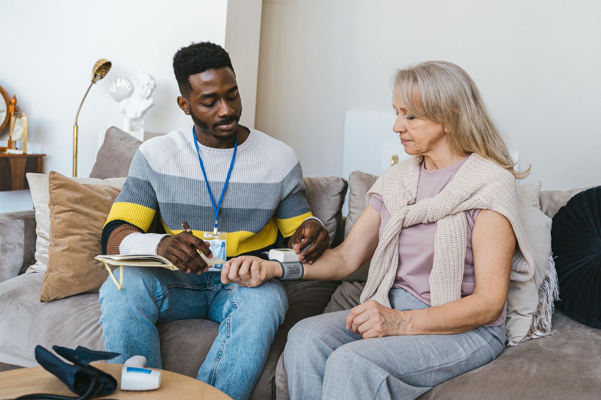 Man takes woman’s blood pressure with a cuff on her arm, both sitting on a sofa in a living room.