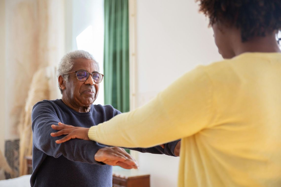 Older adult doing arm exercises with help from a caregiver in a room.