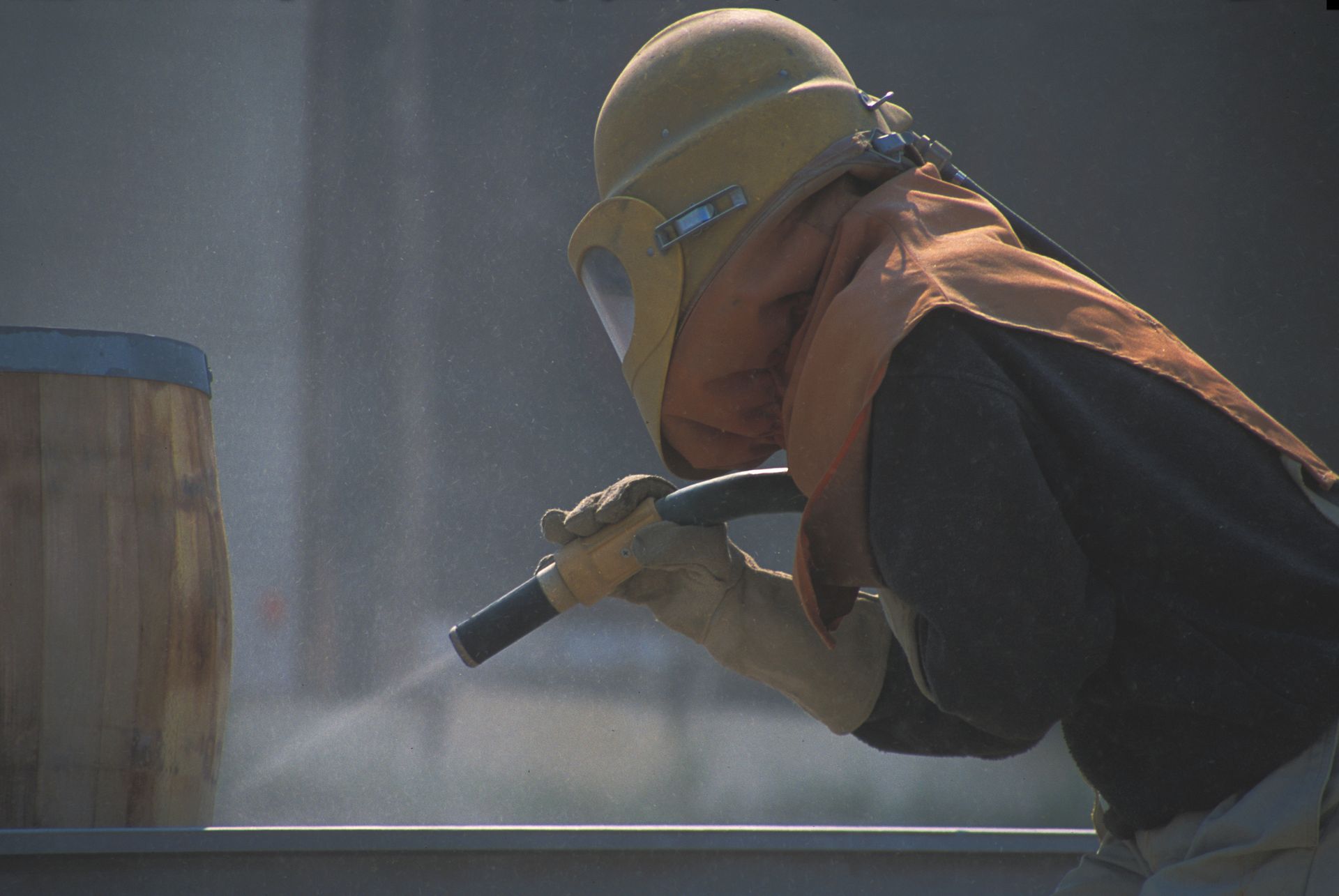 A man wearing a helmet and goggles is sandblasting a wooden barrel.