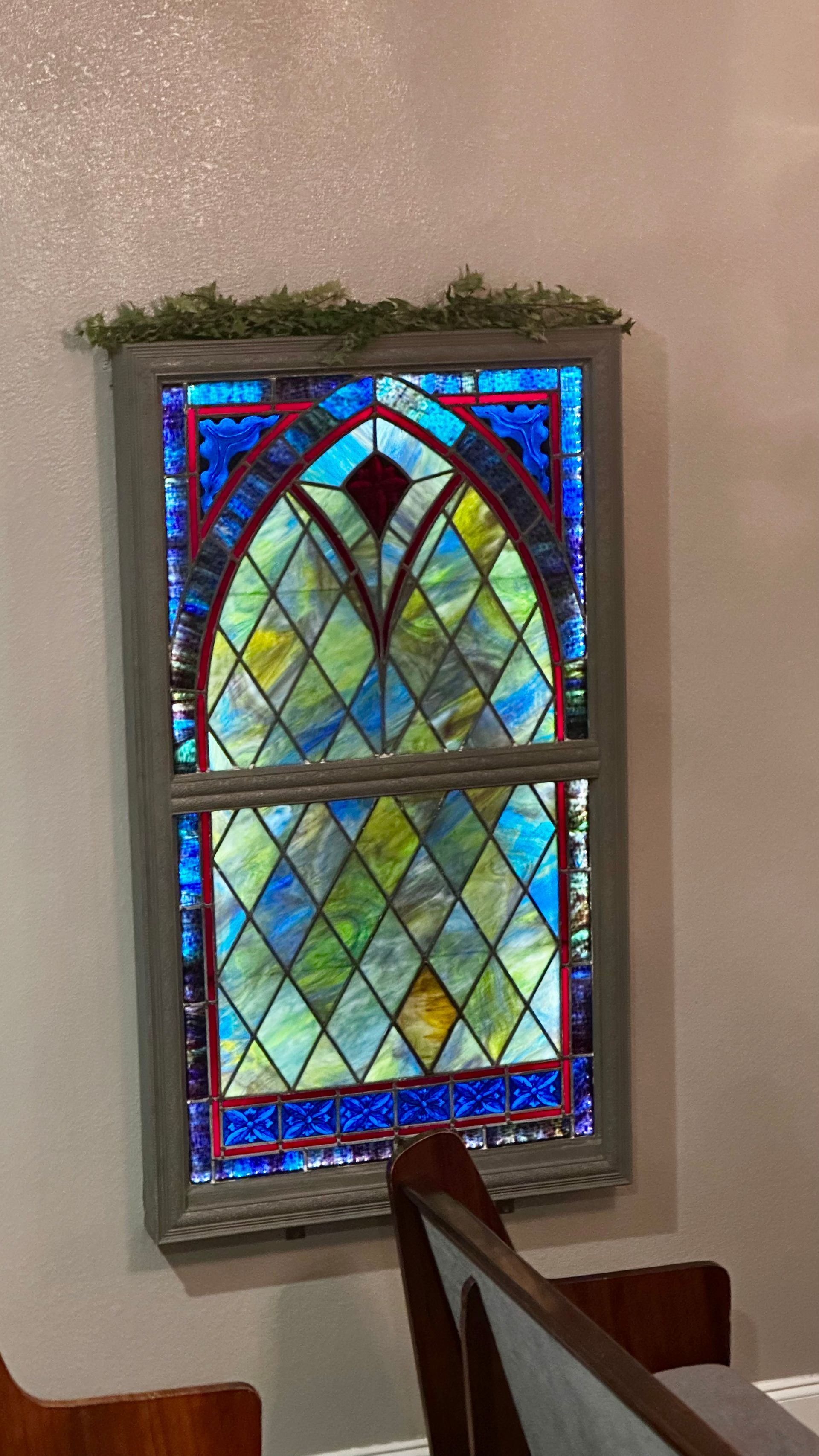 Stained-glass window with blue and red border in a beige wall, partially framed by a chair.