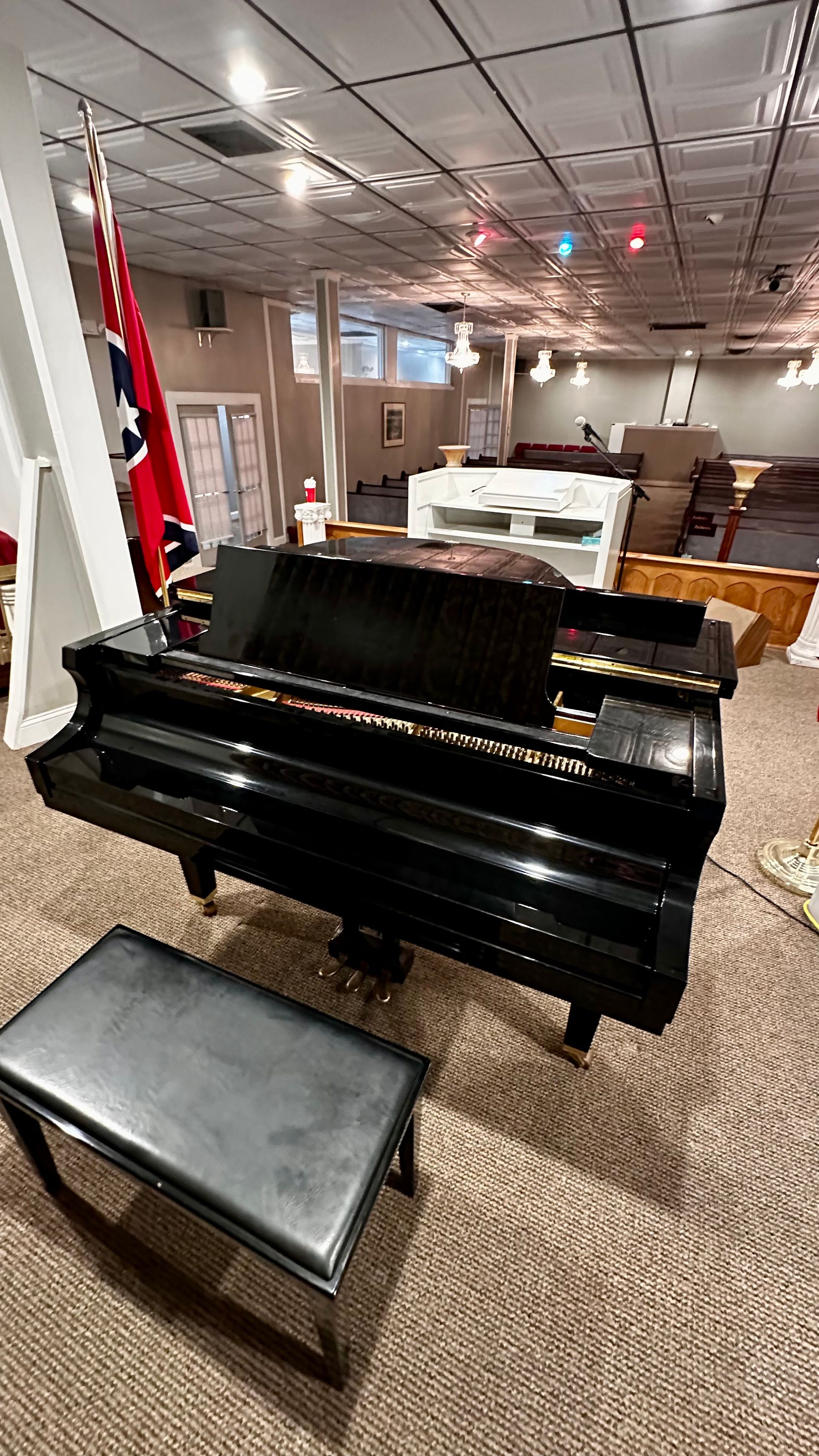 Black grand piano and bench in a carpeted room with flags and tables in the background