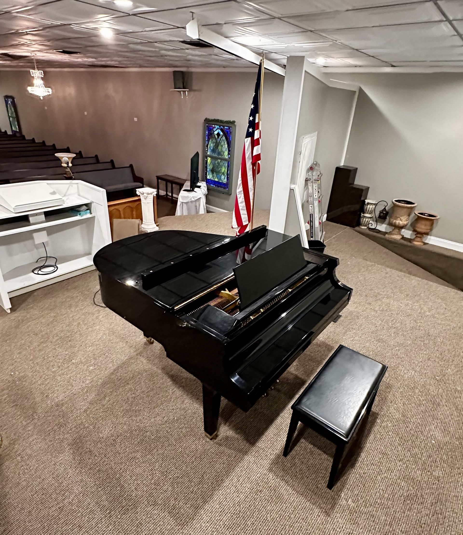 Black grand piano with bench in a carpeted church or hall, near an American flag and seating area