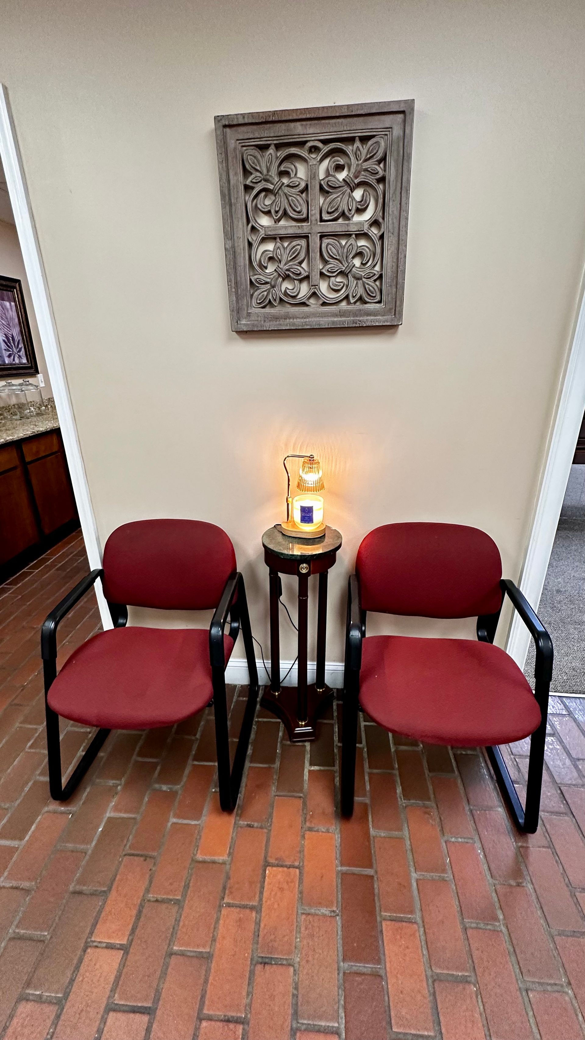 Two red chairs flank a small table with a lamp, against a beige wall and brick floor.