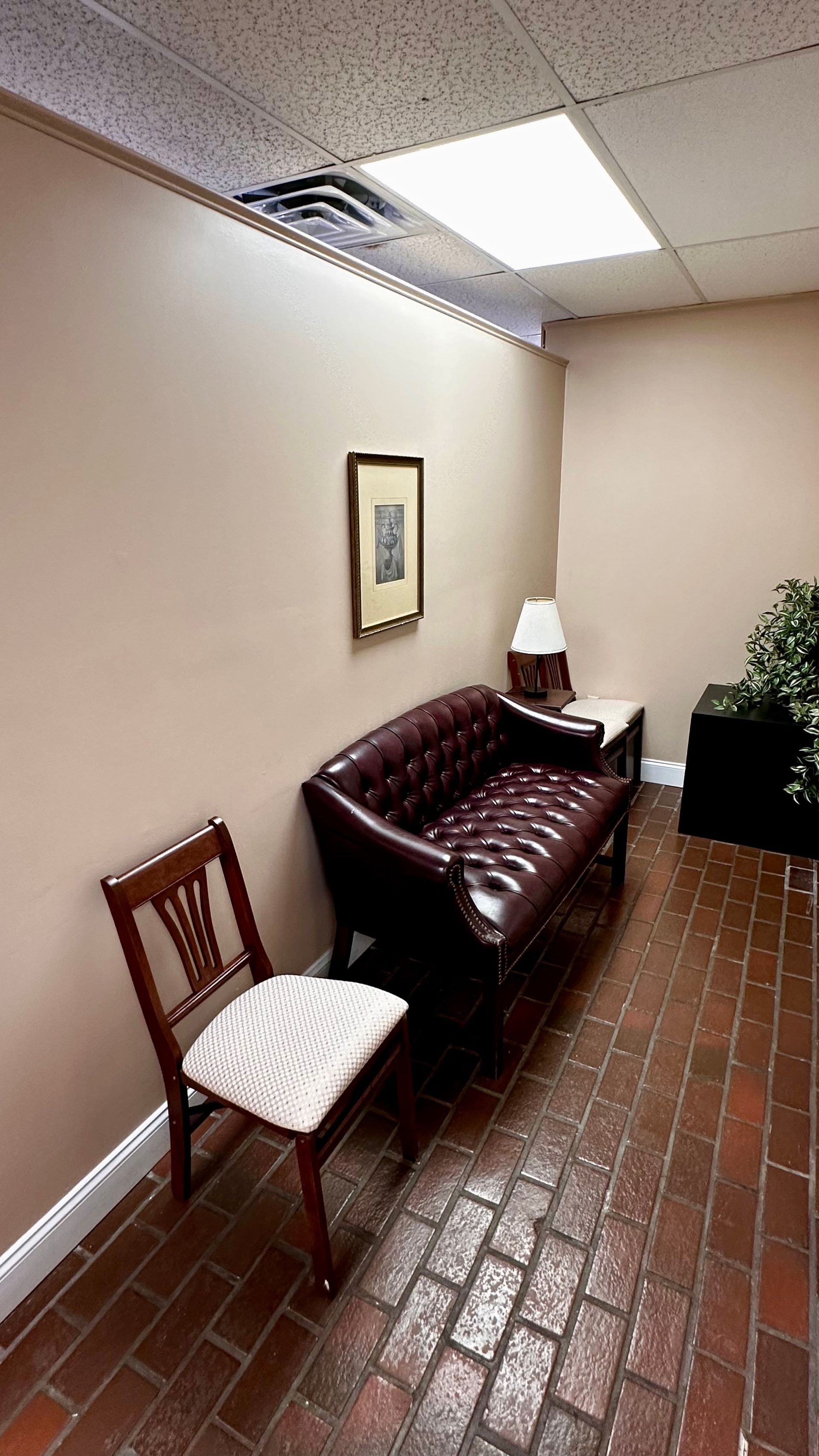 Small waiting area with a brown couch, white chair, framed art, and brick floor under fluorescent lights