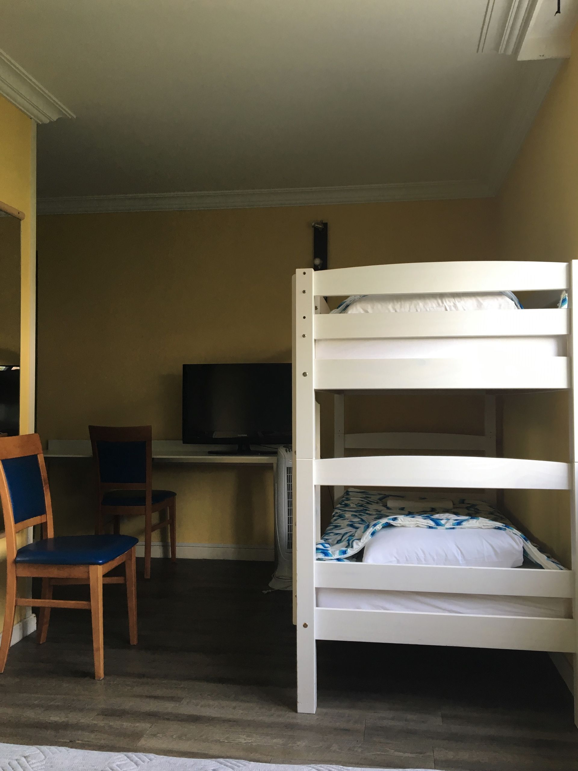 Chambre de dortoir avec lit superposé, bureau, télévision et deux chaises en bois sur un sol foncé
