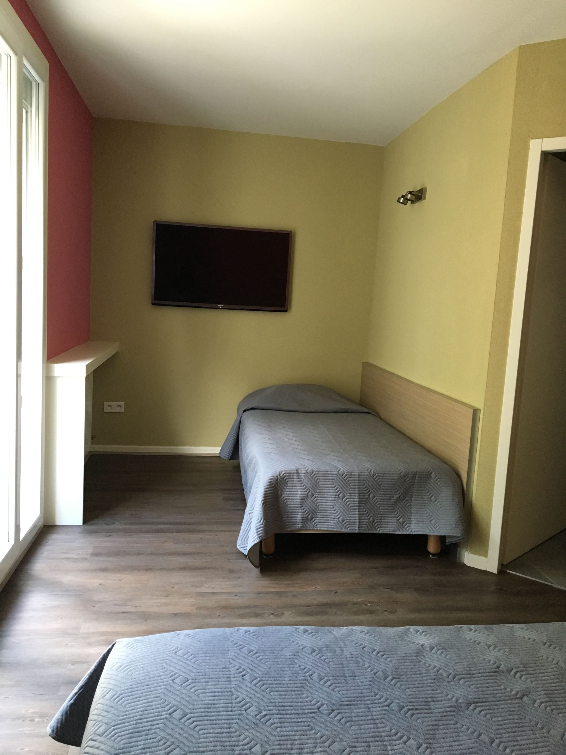 Petite chambre avec deux lits, télévision murale et porte-fenêtre coulissante près d'une fenêtre.