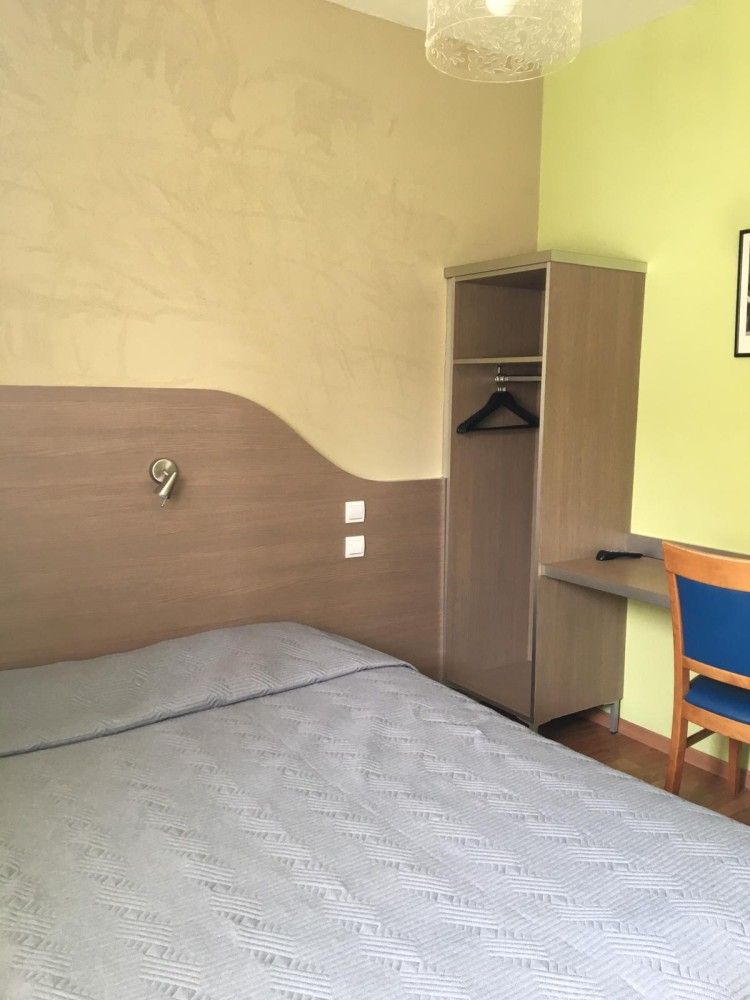 Petite chambre avec lit gris, armoire ouverte et bureau contre un mur jaune.