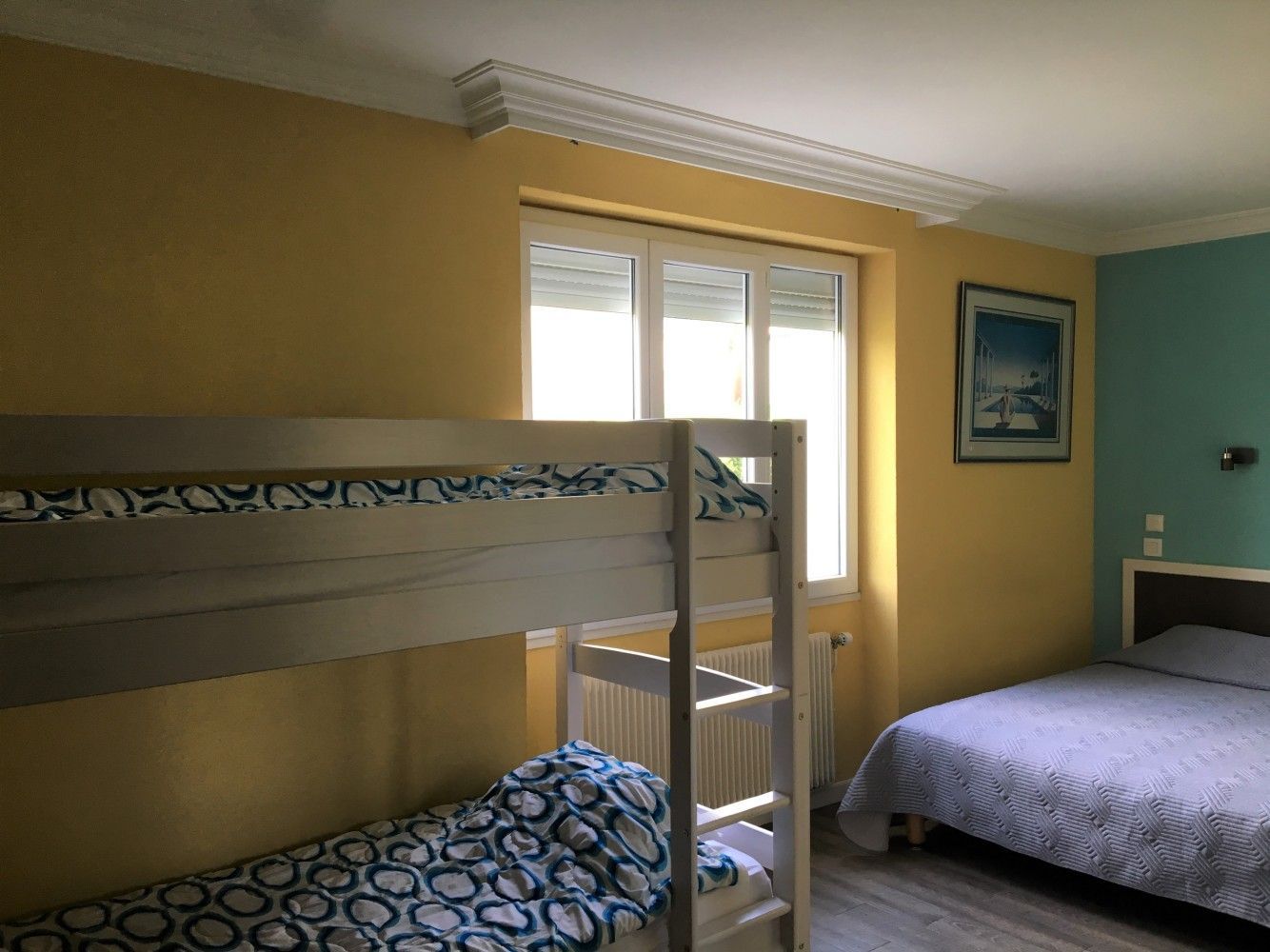 Petite chambre avec lits superposés, murs jaunes et turquoise, et une fenêtre avec stores blancs.