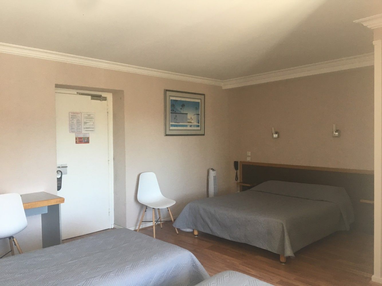 Petite chambre d'hôtel beige avec deux lits, des chaises blanches, un bureau et un tableau encadré au mur.