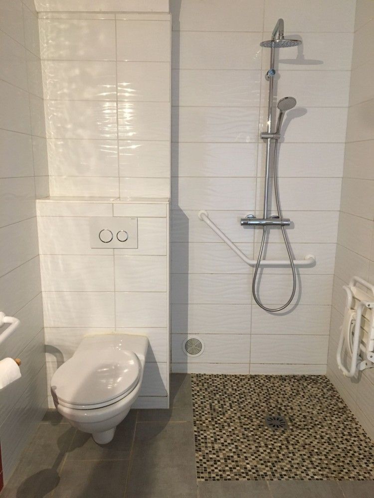 Salle de bain accessible avec toilettes suspendues, douche et barres d'appui sur murs carrelés blancs.