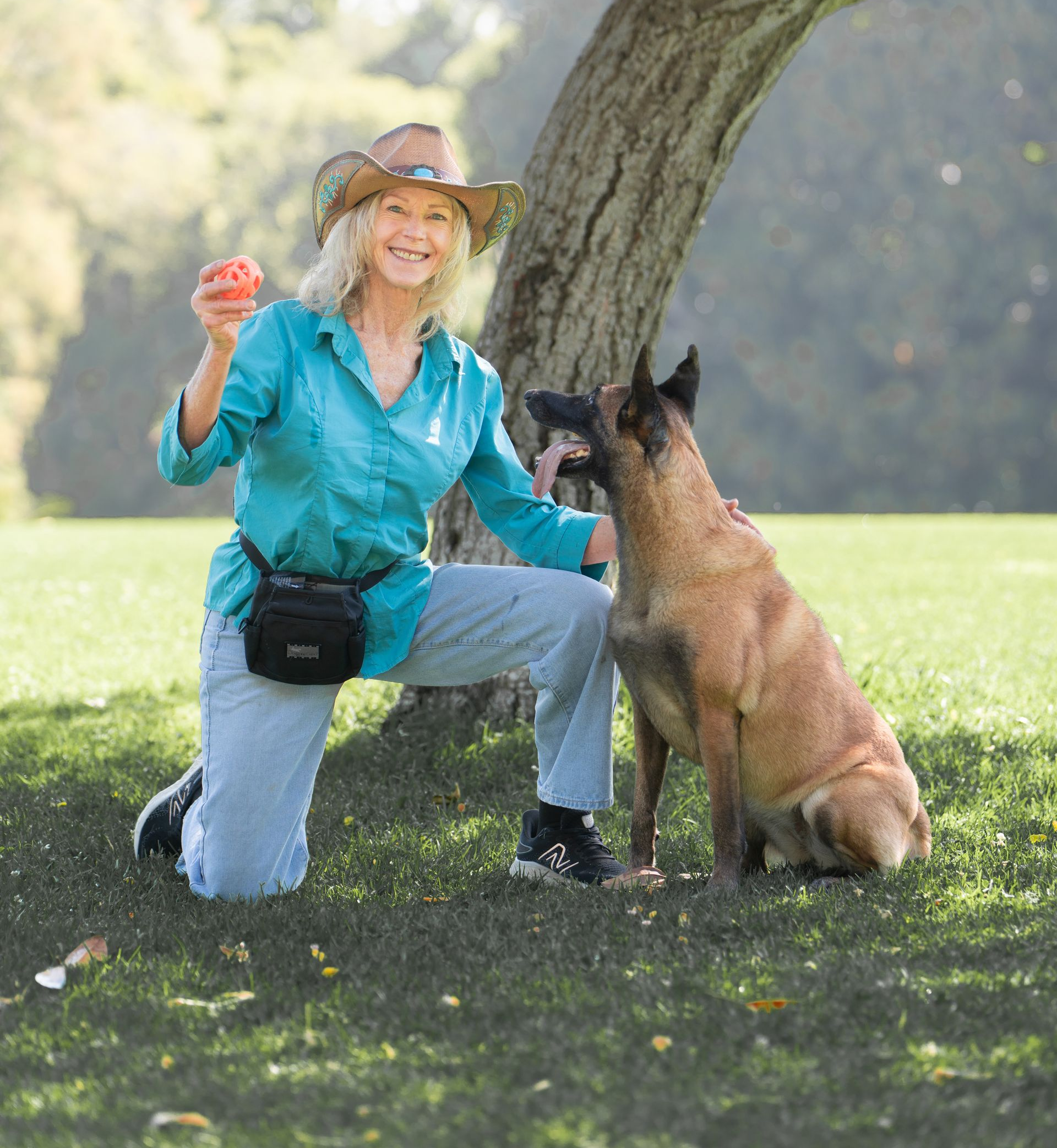 Woman kneeling beside a Belgian Malinois under a tree, holding a red toy in a sunny park.