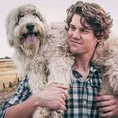 Young man in a plaid shirt carrying a shaggy dog outdoors, both looking content