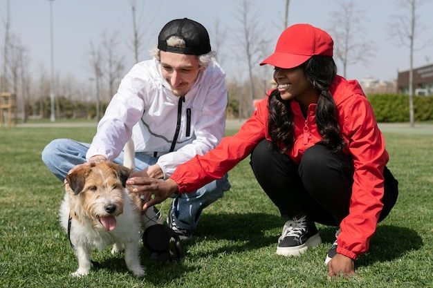 Two people pet a small dog on grass in a park, one in a white hoodie and one in a red jacket and cap.