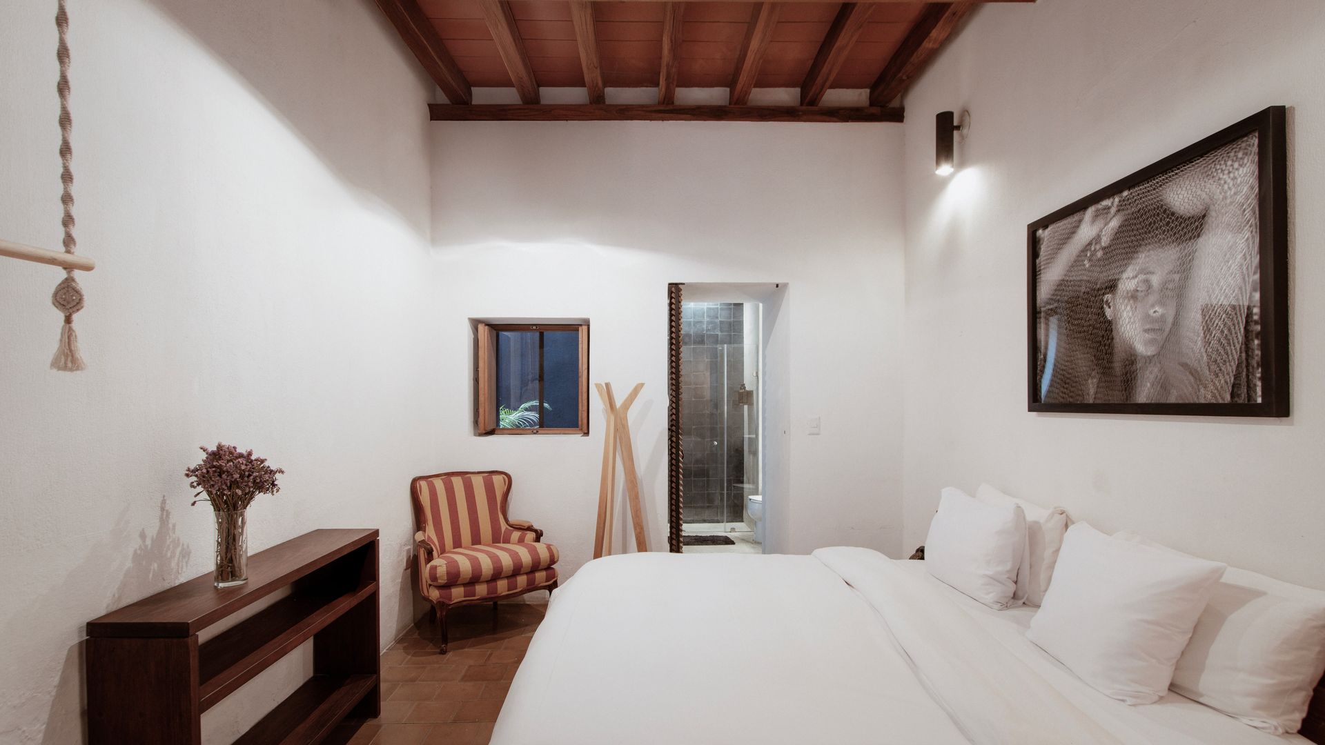 Bright bedroom with a bed, striped armchair, wooden ceiling, and a doorway to a bathroom
