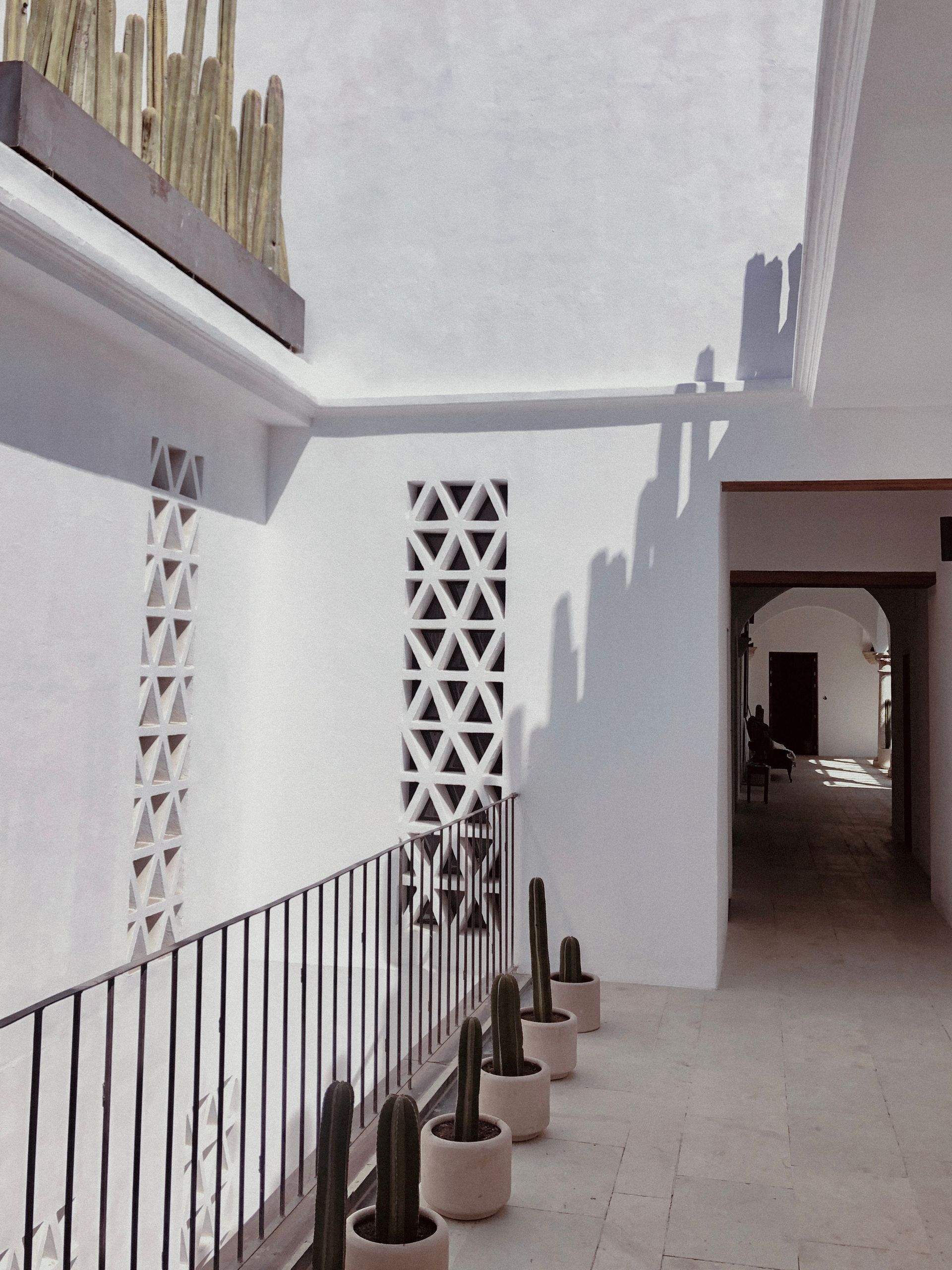 Sunlit white courtyard walkway with geometric lattice windows, railing, and an arched passageway.