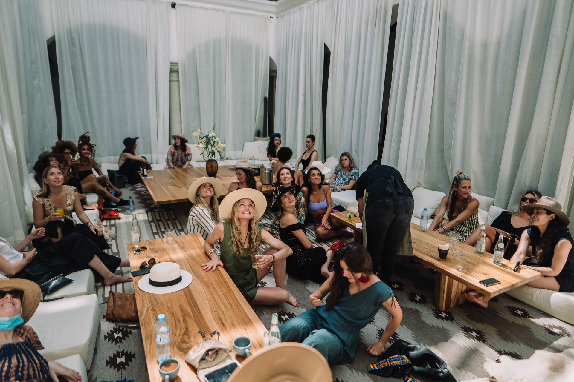 Group of people sitting on low benches around wooden tables in a bright, draped room during a social gathering