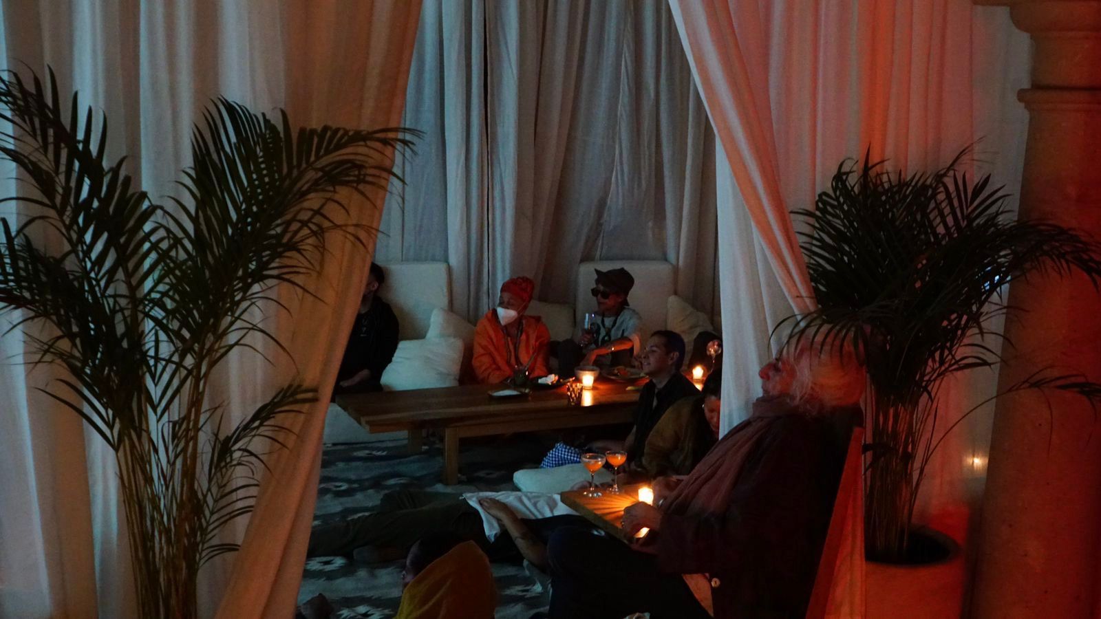 Dimly lit lounge with people seated among curtains and plants, lit by warm candlelight.