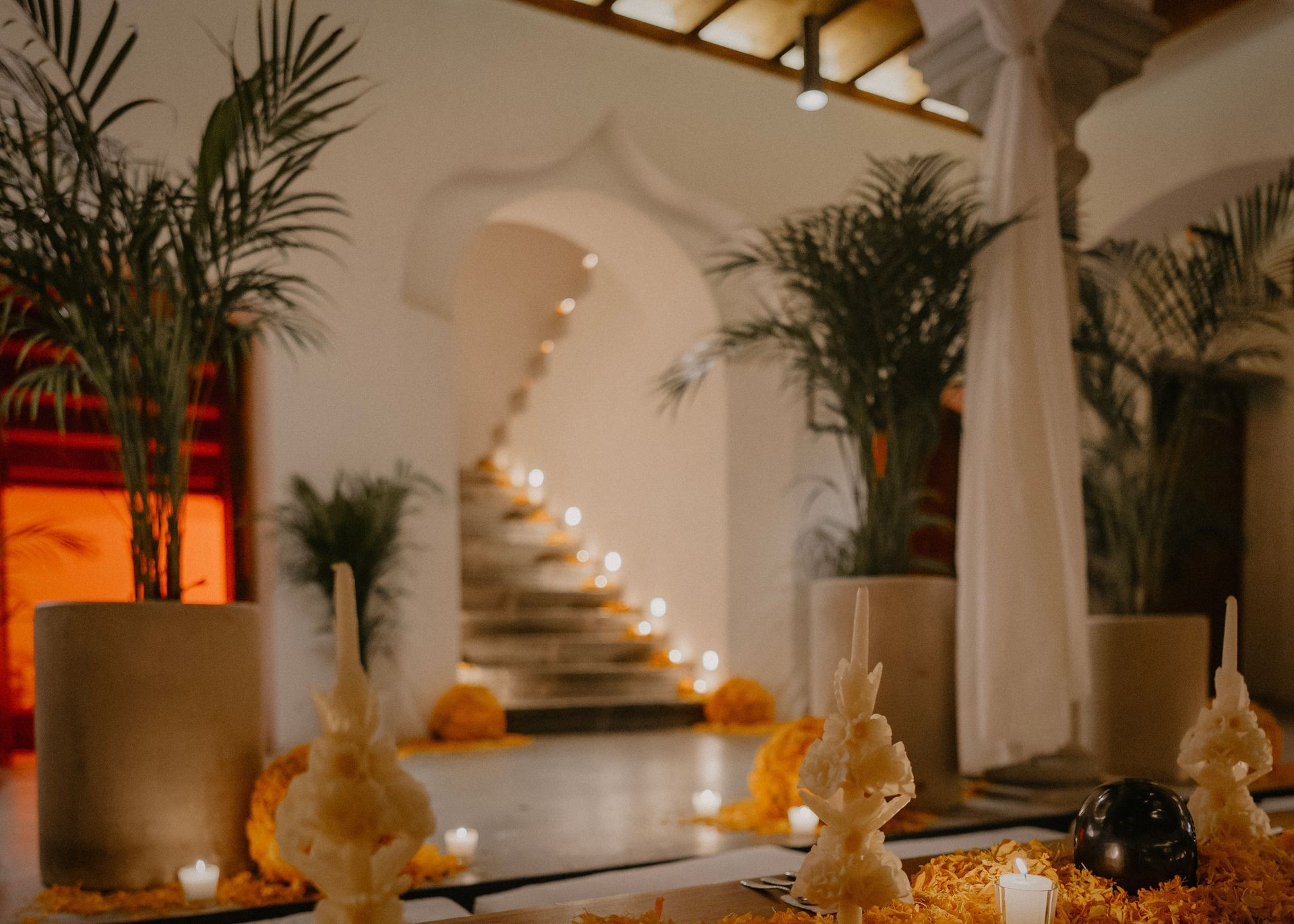 Warm candlelit interior with potted palms, arched stairway, and decorative vases.