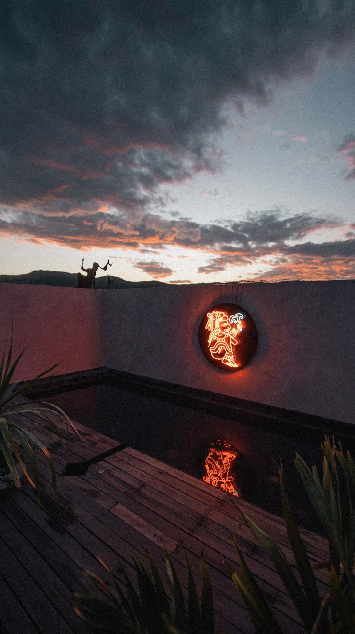 Courtyard pool at dusk beside a white wall with a glowing circular wall light and cloudy pink-blue sky