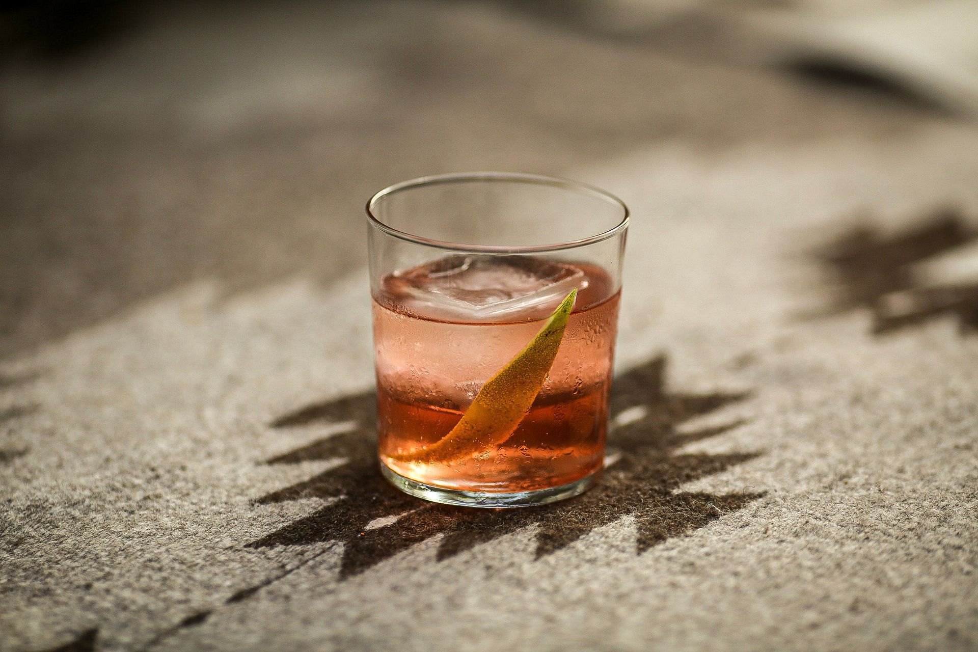A cocktail in a clear glass with ice and an orange peel on a sunlit stone surface.