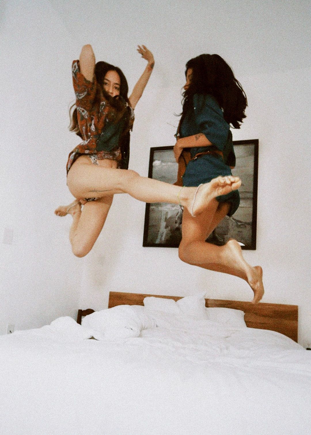Two people mid-jump on a bed in a white bedroom, arms raised and smiling.