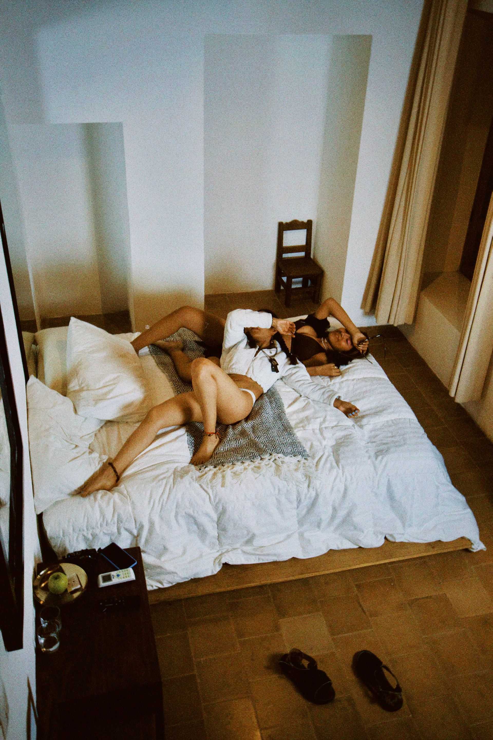 Two people lying on a bed in a sparsely furnished room, with shoes on the floor nearby.