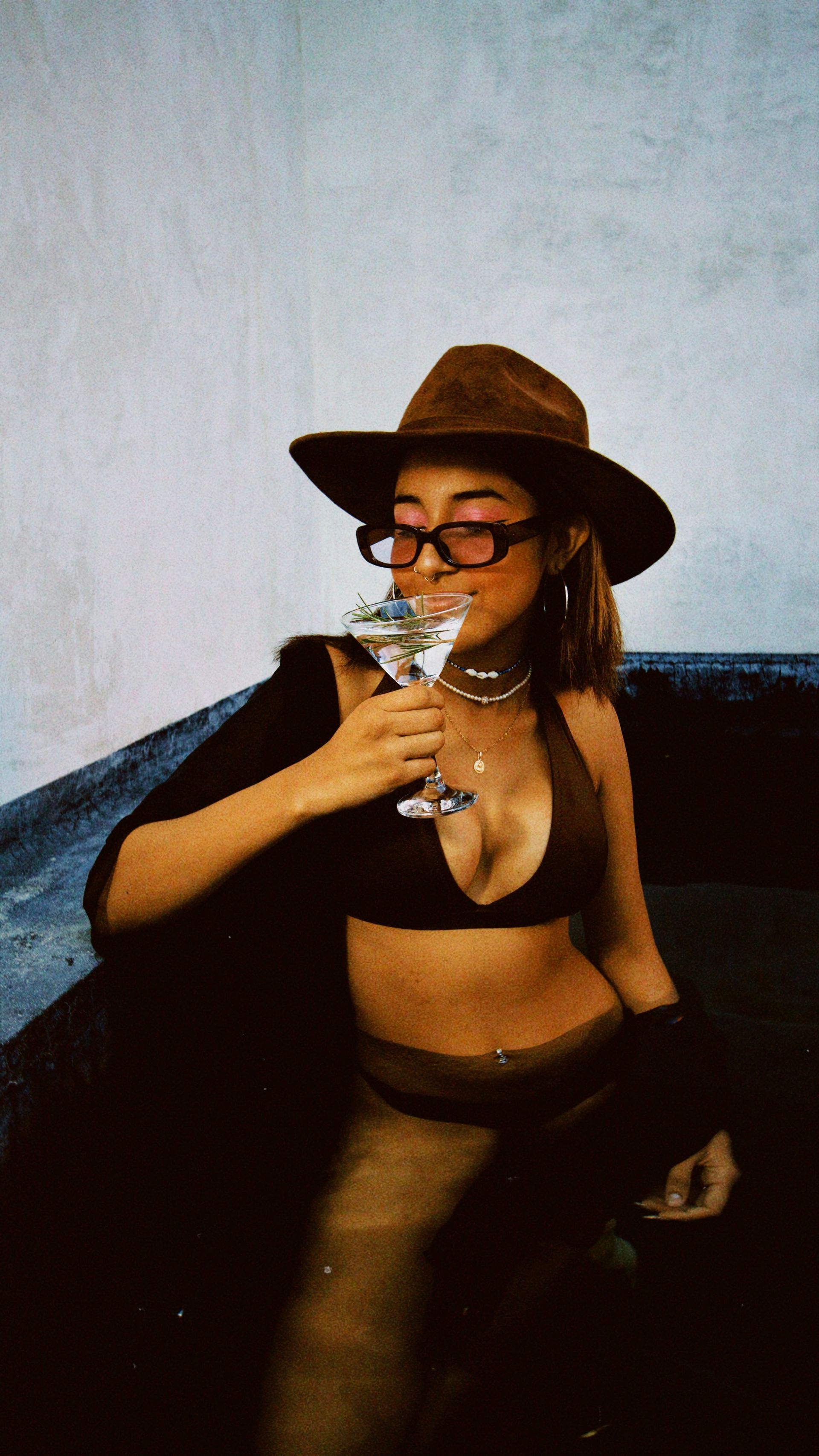 Person in a brown hat and glasses holding a drink while seated on a dark couch.
