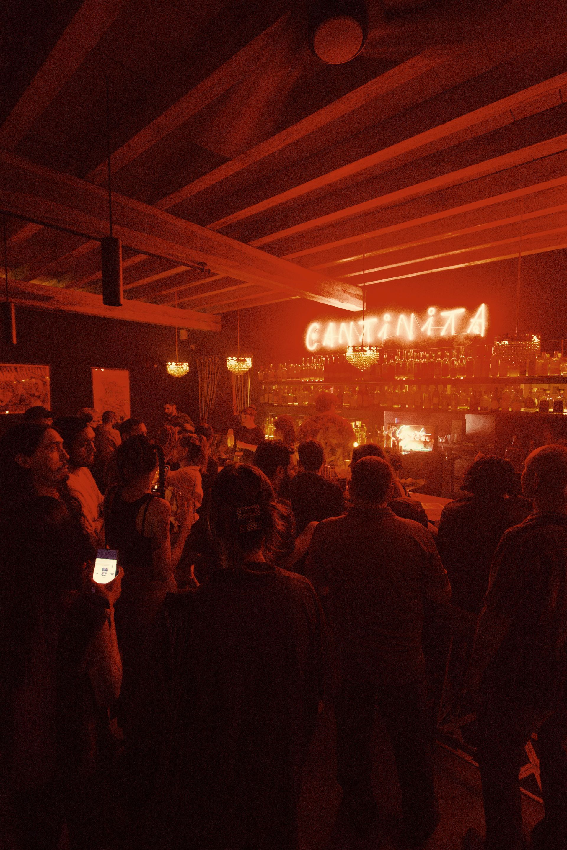 Crowd gathered in a dim red-lit venue with bright lights over a stage and exposed ceiling beams