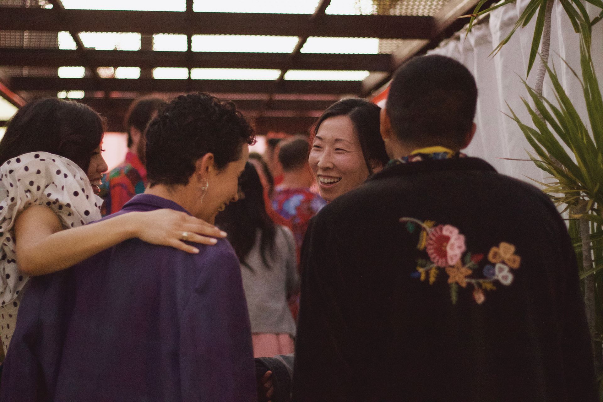 Friends chatting and hugging at an outdoor gathering under a wooden pergola.