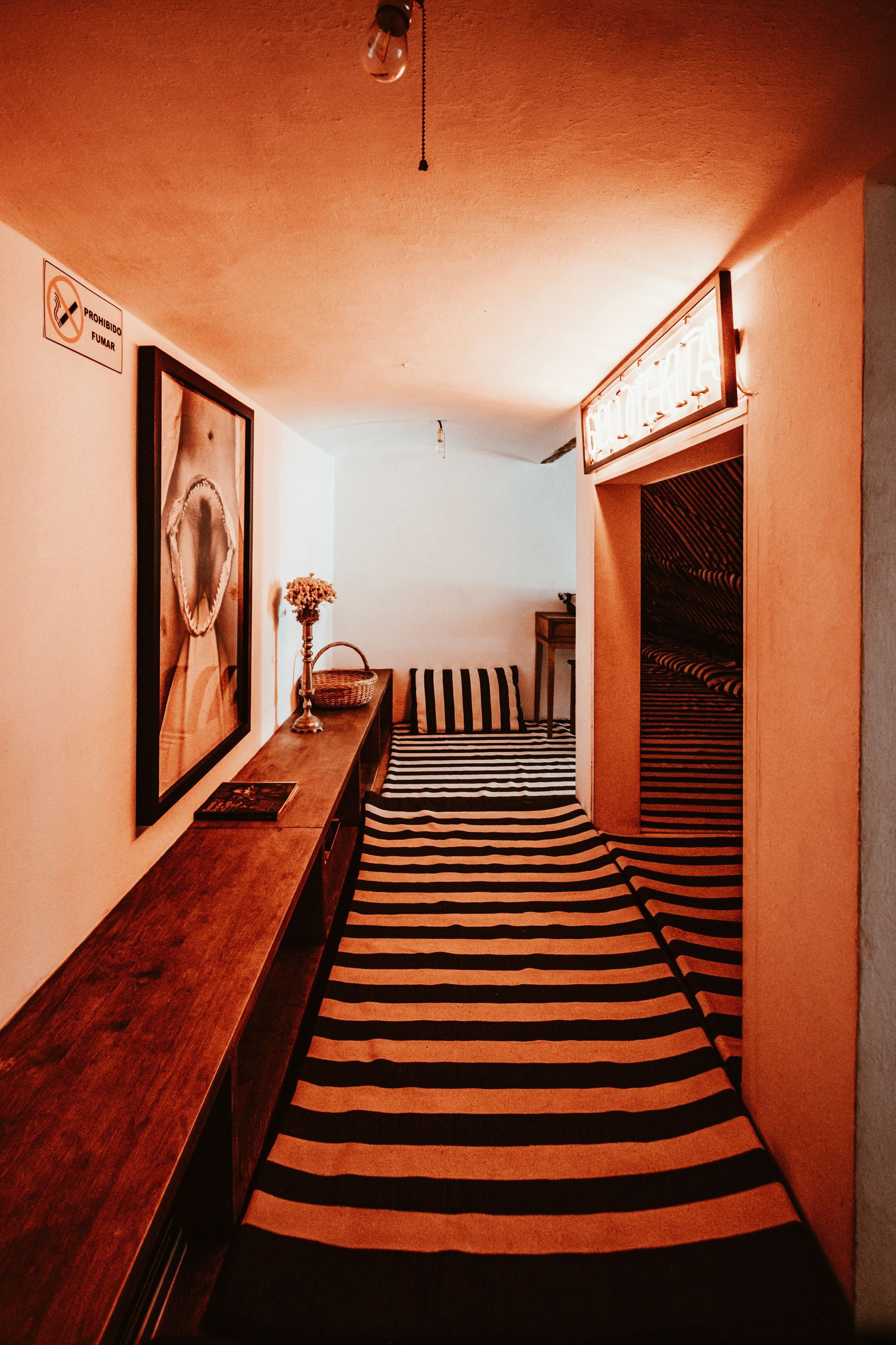 Warmly lit hallway with striped carpet, framed art, and a narrow stairway at the end.